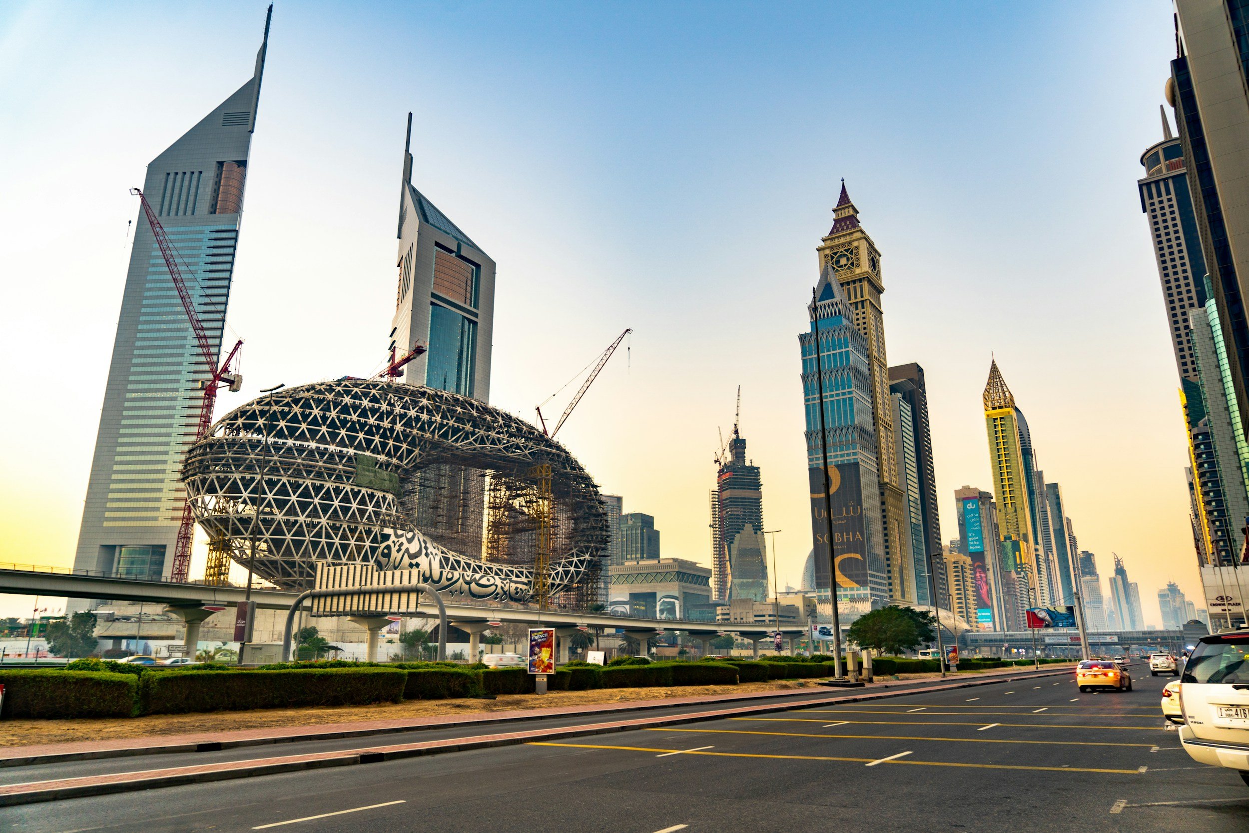Cityscape of Dubai with skyscrapers including the Cayan Tower, and the Deira City Center shopping mall under construction, with a street and cars in the foreground.