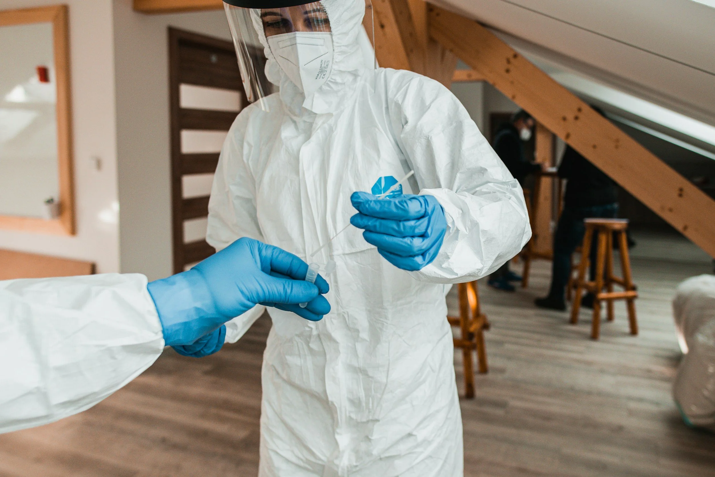 A healthcare worker wearing full personal protective equipment, including a face shield, mask, white gown, and blue gloves, is handling a swab in an indoor setting.
