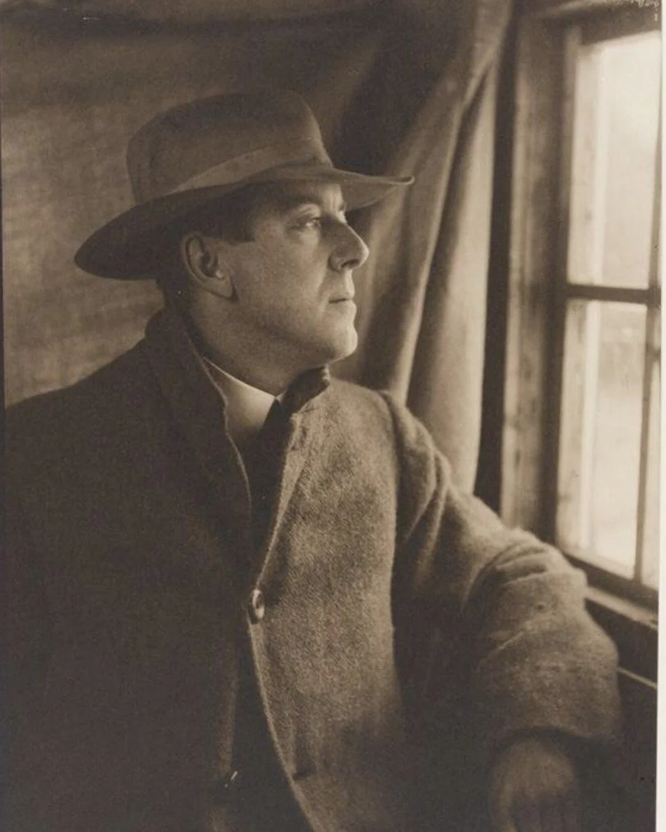 A black-and-white photograph of a man wearing a hat and a wool coat, sitting by a window and looking outside thoughtfully.