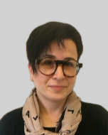 A person with short dark hair wearing large sunglasses, a beige scarf with black patterns, and a black top, against a plain background.