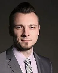 A young man with light skin, short dark hair, wearing a dark suit, white shirt, and patterned tie, posed against a dark background.