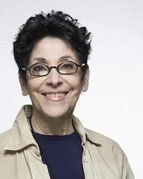 Smiling middle-aged woman with short dark curly hair and glasses, wearing a beige jacket over a dark top, against a white background.