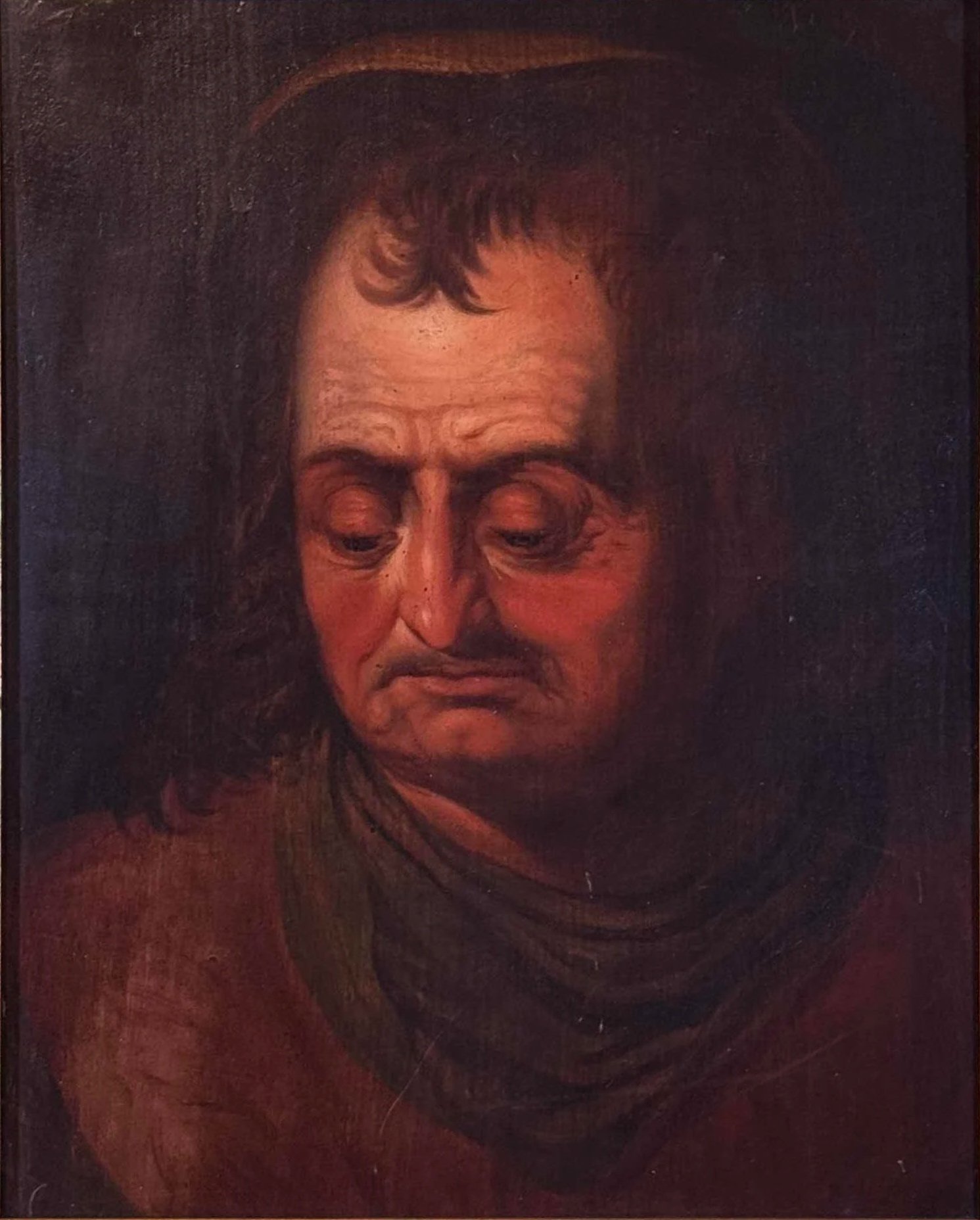 A close-up portrait of a man with long, curly hair, a frowning expression, and wearing a brown coat, set against a dark background.