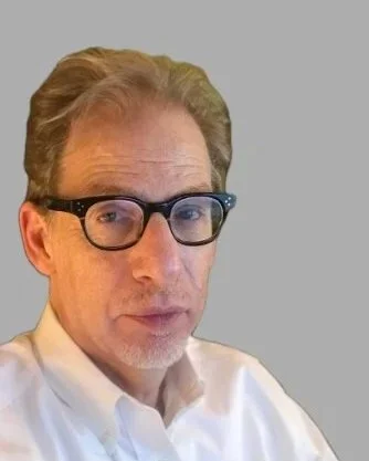 Close-up of a middle-aged man with light-colored hair, glasses, and a white collared shirt against a plain background.
