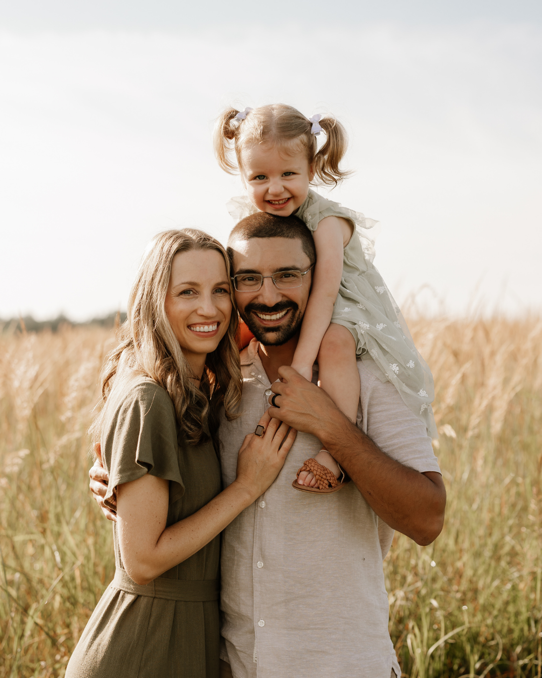 mom, dad, and little girl sitting on dads shoulders, smiling at the camera in a tall field of grass, styled for family photos