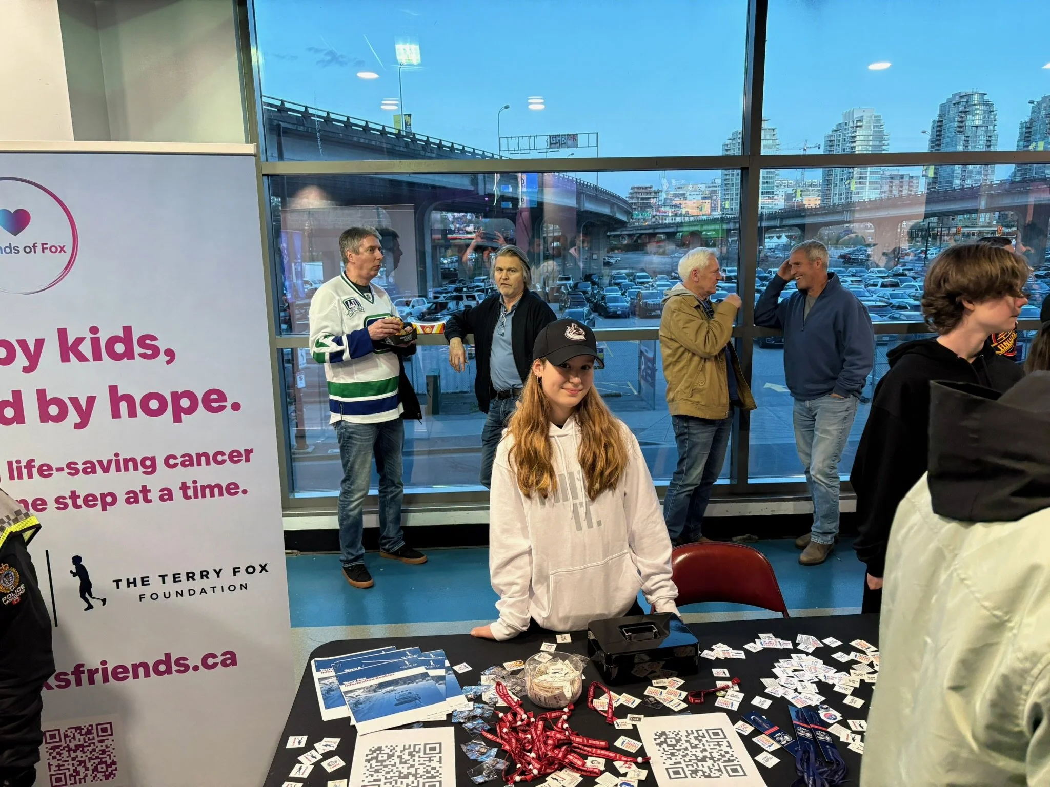 A group of people are gathered inside at an event, with some standing near a table covered with brochures, lanyards, and QR codes. A young girl with long red hair, wearing a black cap and white hoodie, is seated at the table, smiling at the camera. B