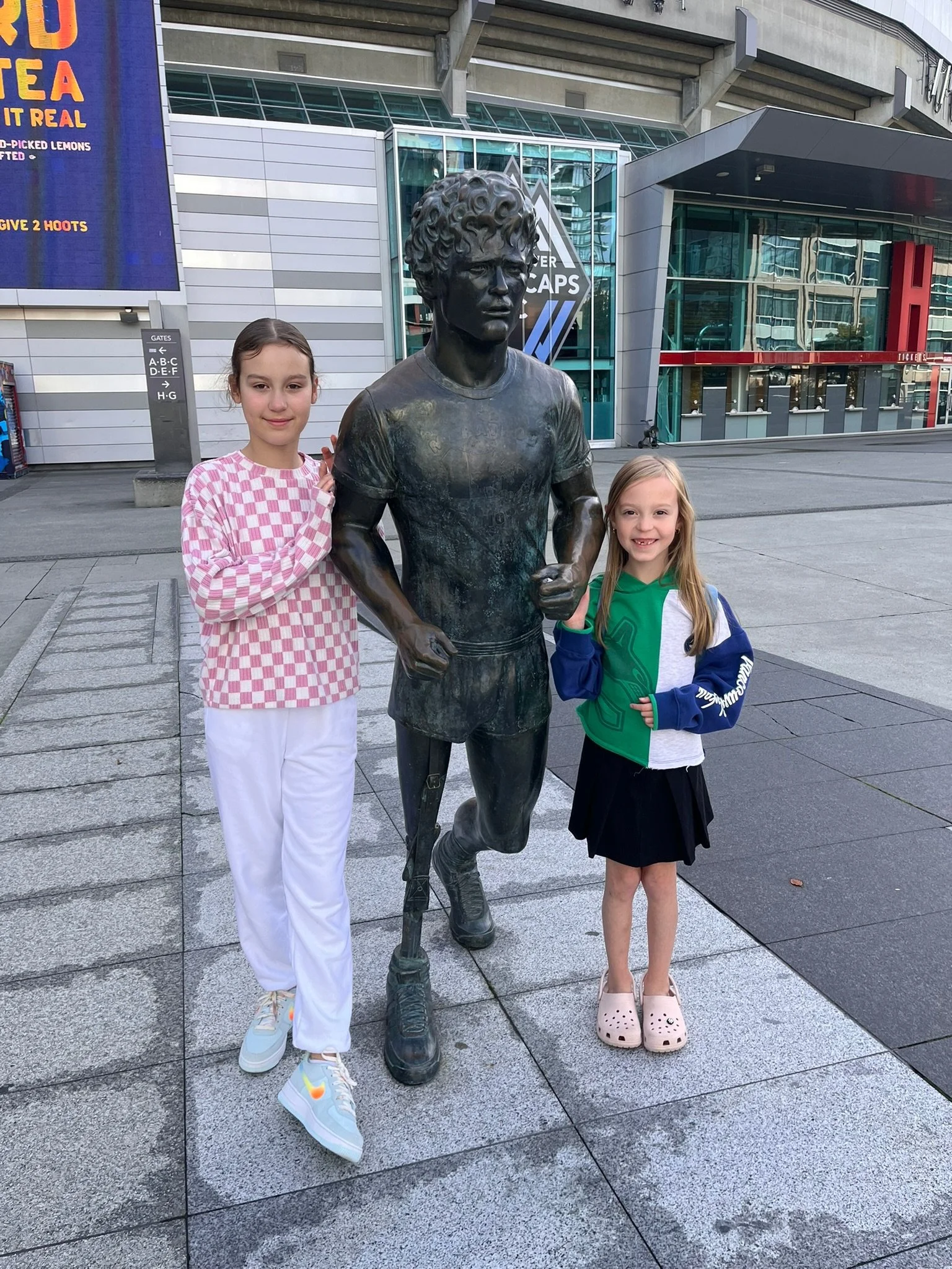 Two young girls standing beside a statue of a male athlete. The girl on the left has brown hair in a ponytail, wearing a pink and white checkered long-sleeve shirt and white pants. The girl on the right has blonde hair, wearing a green, white, and bl