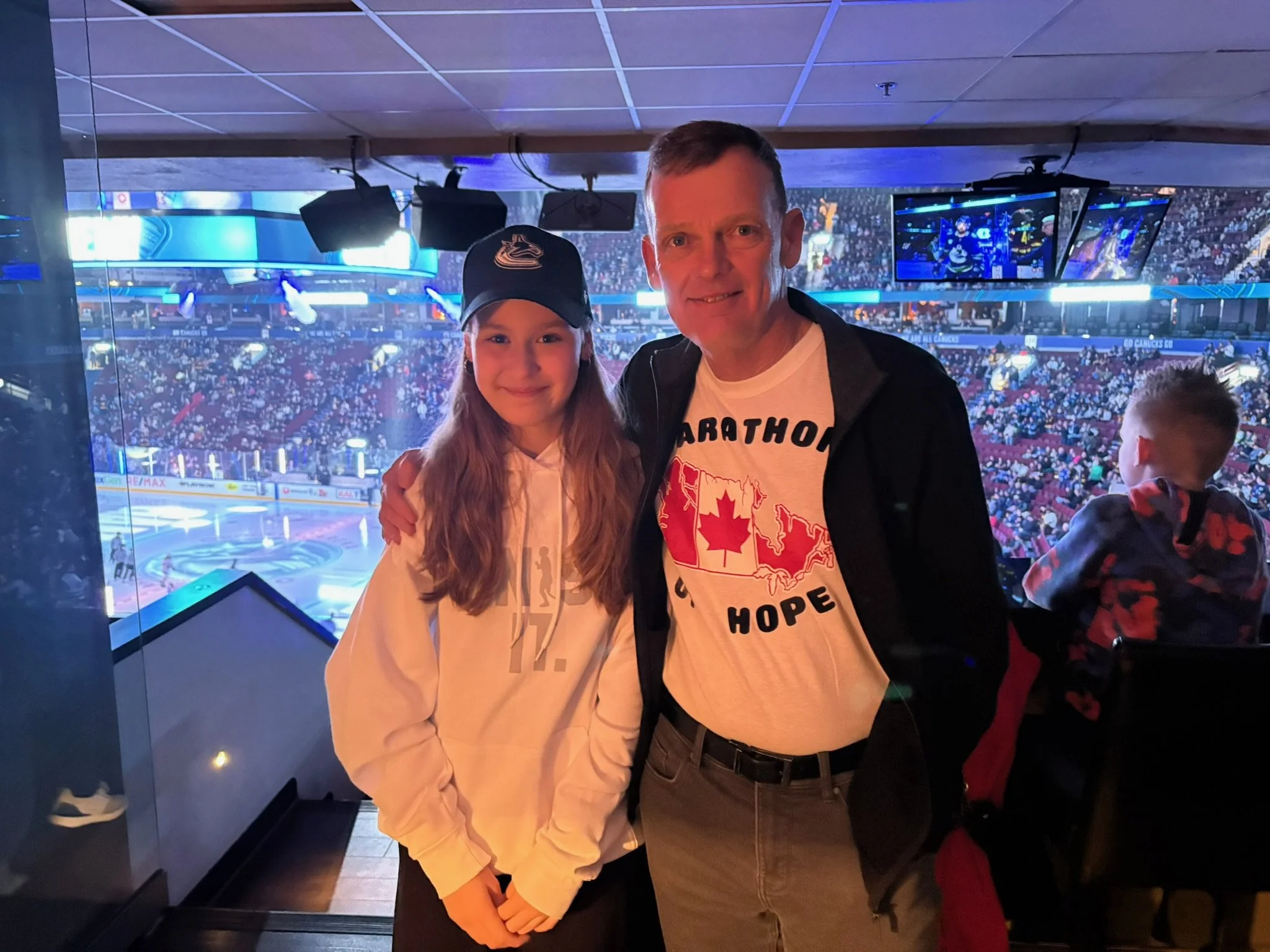 A girl and an older man standing together at an indoor ice hockey game, with the rink and audience in the background. The girl is wearing a white hoodie and a black baseball cap, and the man is wearing a white T-shirt with a Canadian map and the words 'Marathon for Hope.' They are smiling and posing for the photo.
