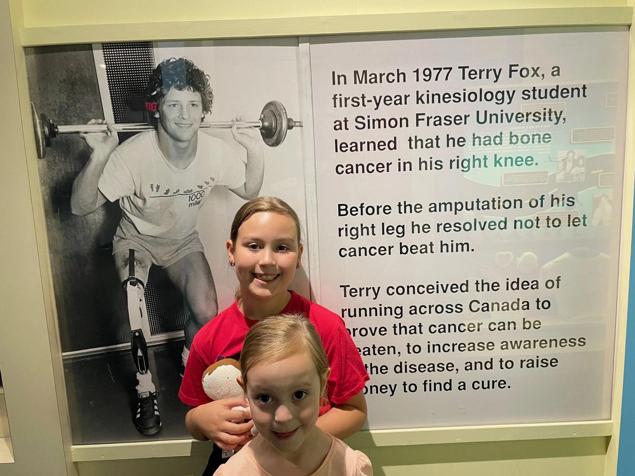Two young girls smiling and standing in front of a large black-and-white poster of Terry Fox, a cancer survivor and runner, with a younger girl holding a donut in her hand. The poster includes a photograph of Terry Fox lifting weights and text about his story and the idea of running across Canada to raise awareness and funds for cancer research.