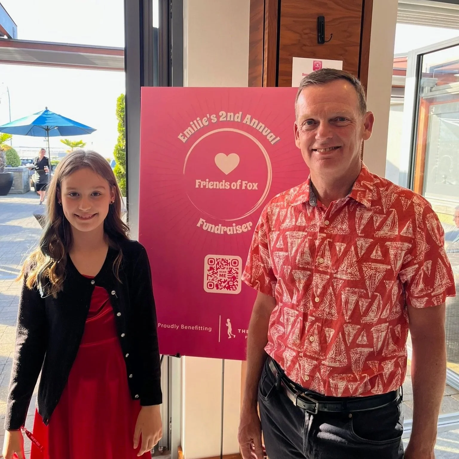 A young girl and an adult man smiling and standing beside a pink poster that reads 'Emilie’s 2nd Annual Friends of Fox Fundraiser' with a QR code, inside a building with glass doors and an outdoor patio visible in the background.