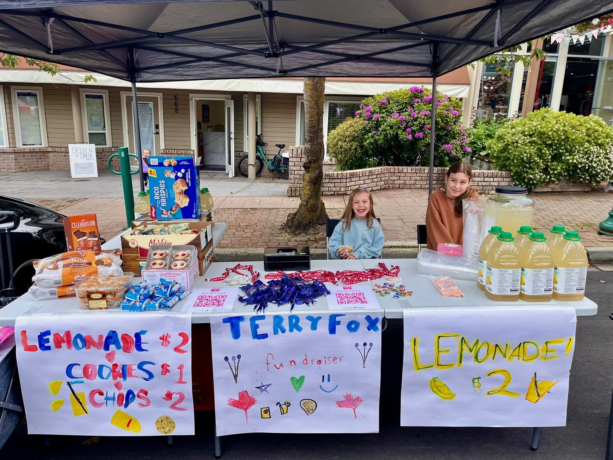 A lemonade stand with two young girls selling lemonade and baked goods outdoors on a street, under a canopy, with colorful signs advertising items and prices.