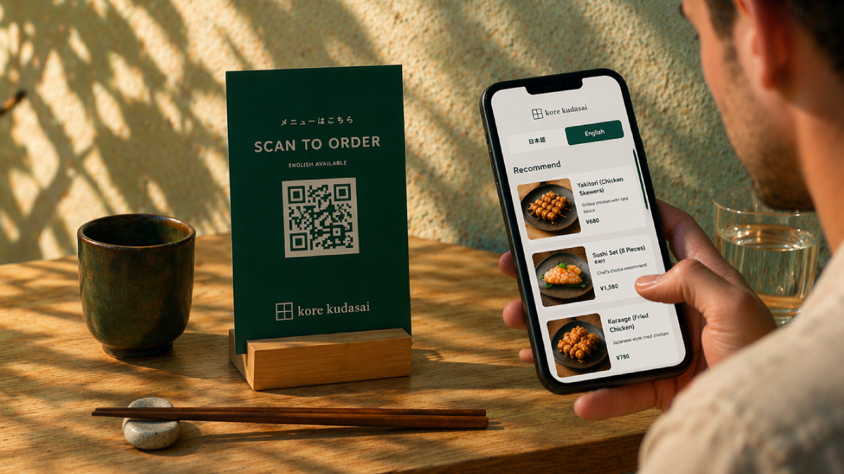 A person holds a smartphone displaying a digital menu with Japanese and English options, featuring sushi and fried chicken dishes. The table has a green QR code sign for ordering, a ceramic cup, a glass of water, and chopsticks with a chopstick rest.