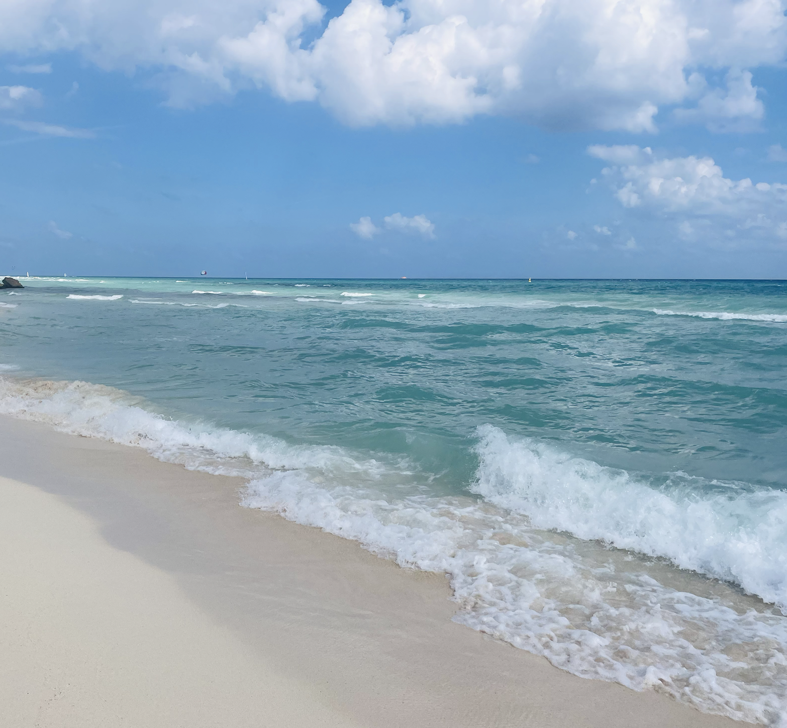 Sunny beach with clear turquoise water, white sandy shore, and blue sky with scattered clouds.