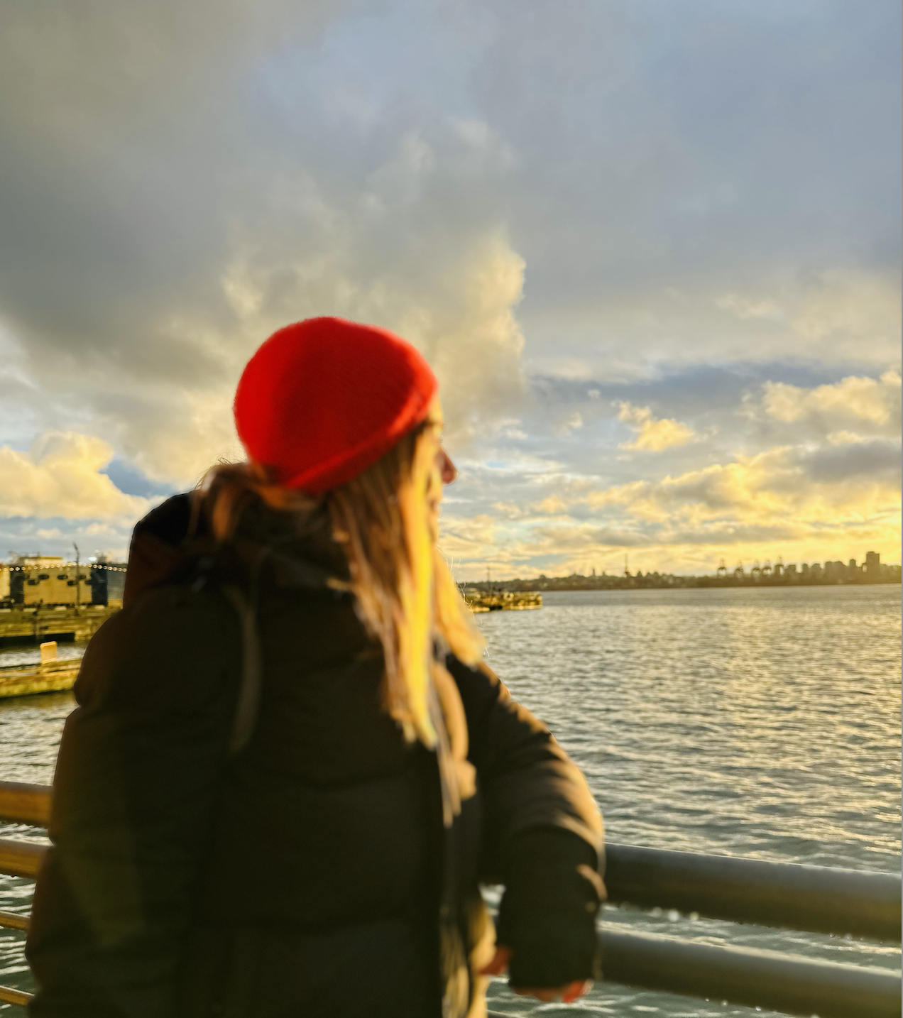 A woman with long hair wearing a red beanie and sunglasses is leaning on a railing on a boat or dock, looking towards the water during sunset with cityscape in the distance.