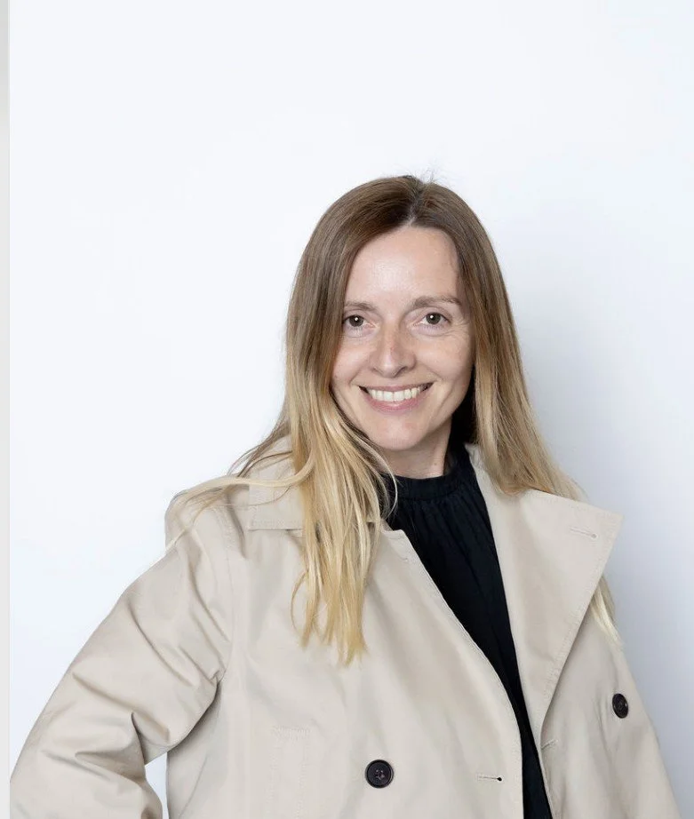 A woman with long blonde hair smiling, wearing a beige trench coat over a black top, standing against a plain white background.
