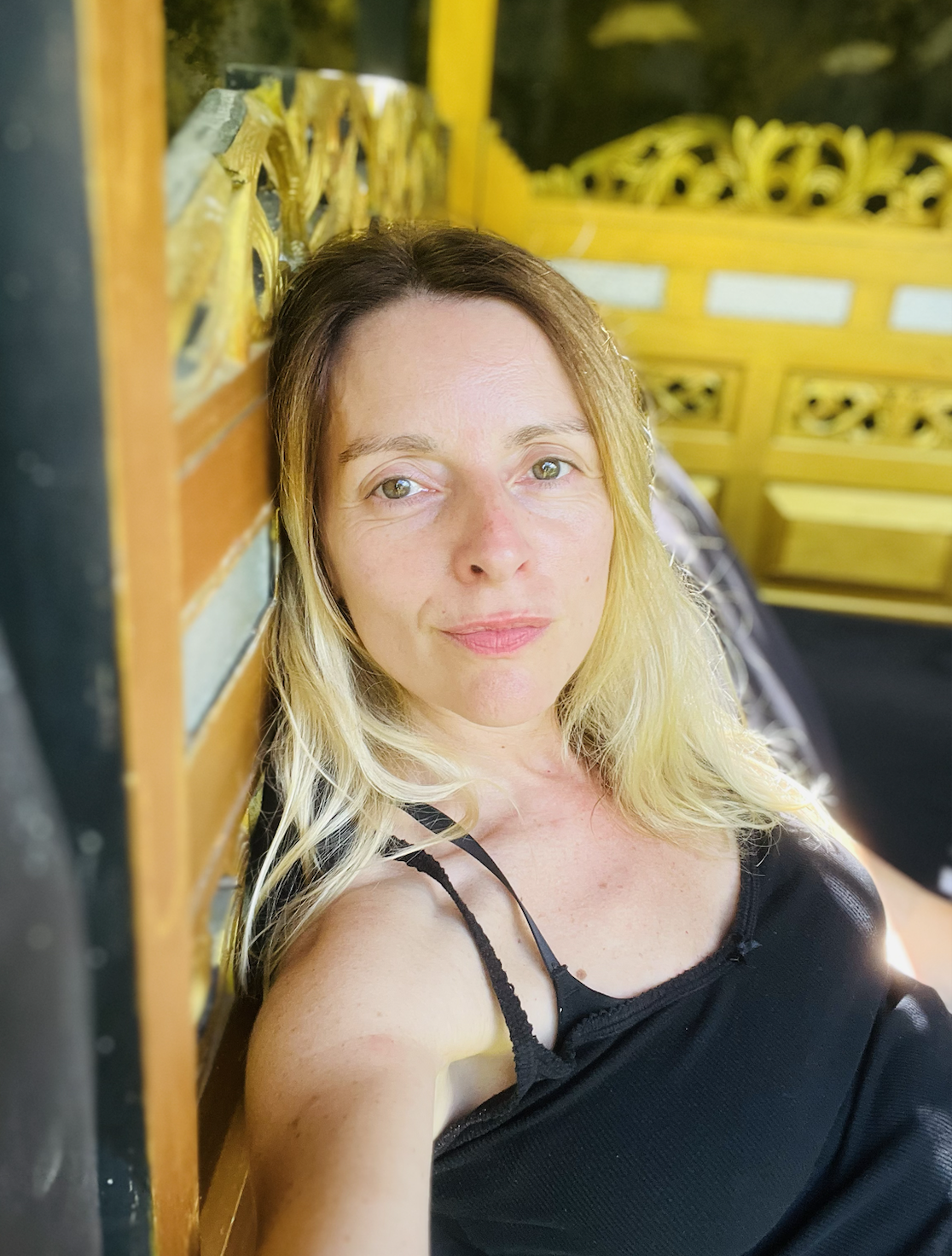 A woman with blonde and brown hair lying on a yellow decorative bench, wearing a black spaghetti strap top, outdoors during daytime.