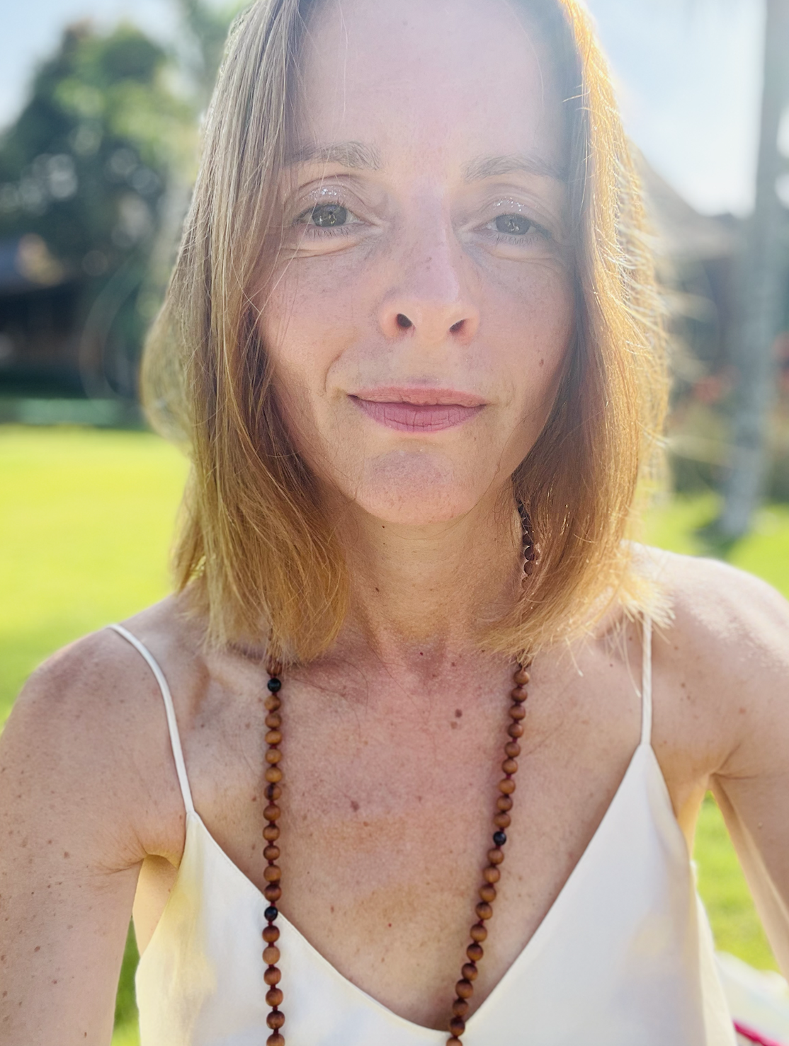 Close-up of a woman with shoulder-length light brown hair, wearing a white spaghetti strap top and a beaded necklace, smiling outdoors in bright sunlight with a blurred green background.