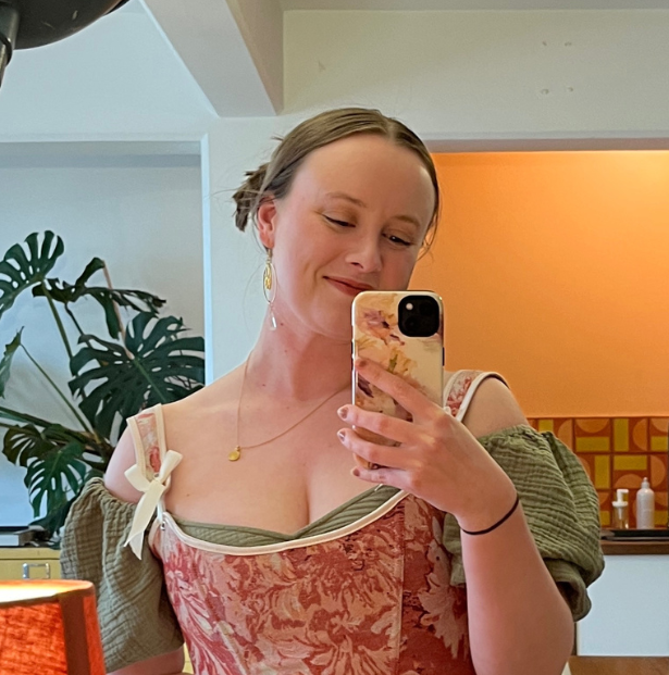 A woman taking a mirror selfie with her phone, smiling slightly, wearing a floral dress and earrings, in a warmly lit room with plants and abstract art in the background.