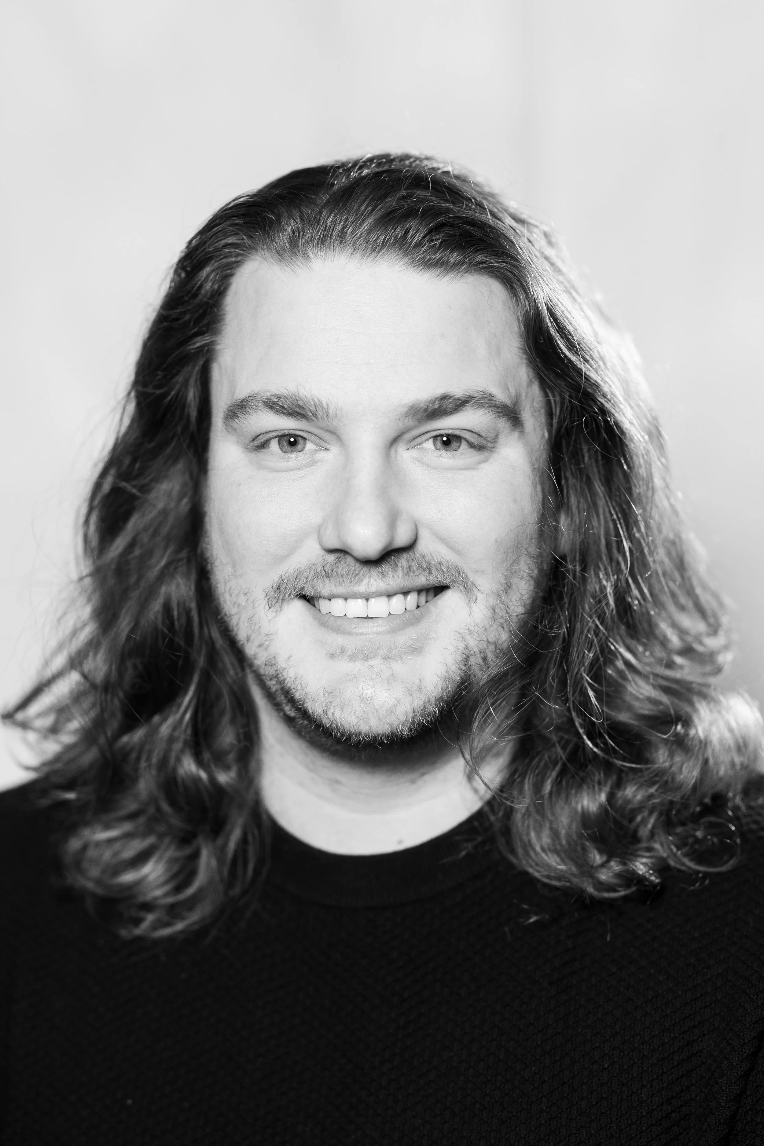 Black and white portrait of a smiling man with long, wavy hair and a beard, wearing a black shirt, looking directly at the camera.