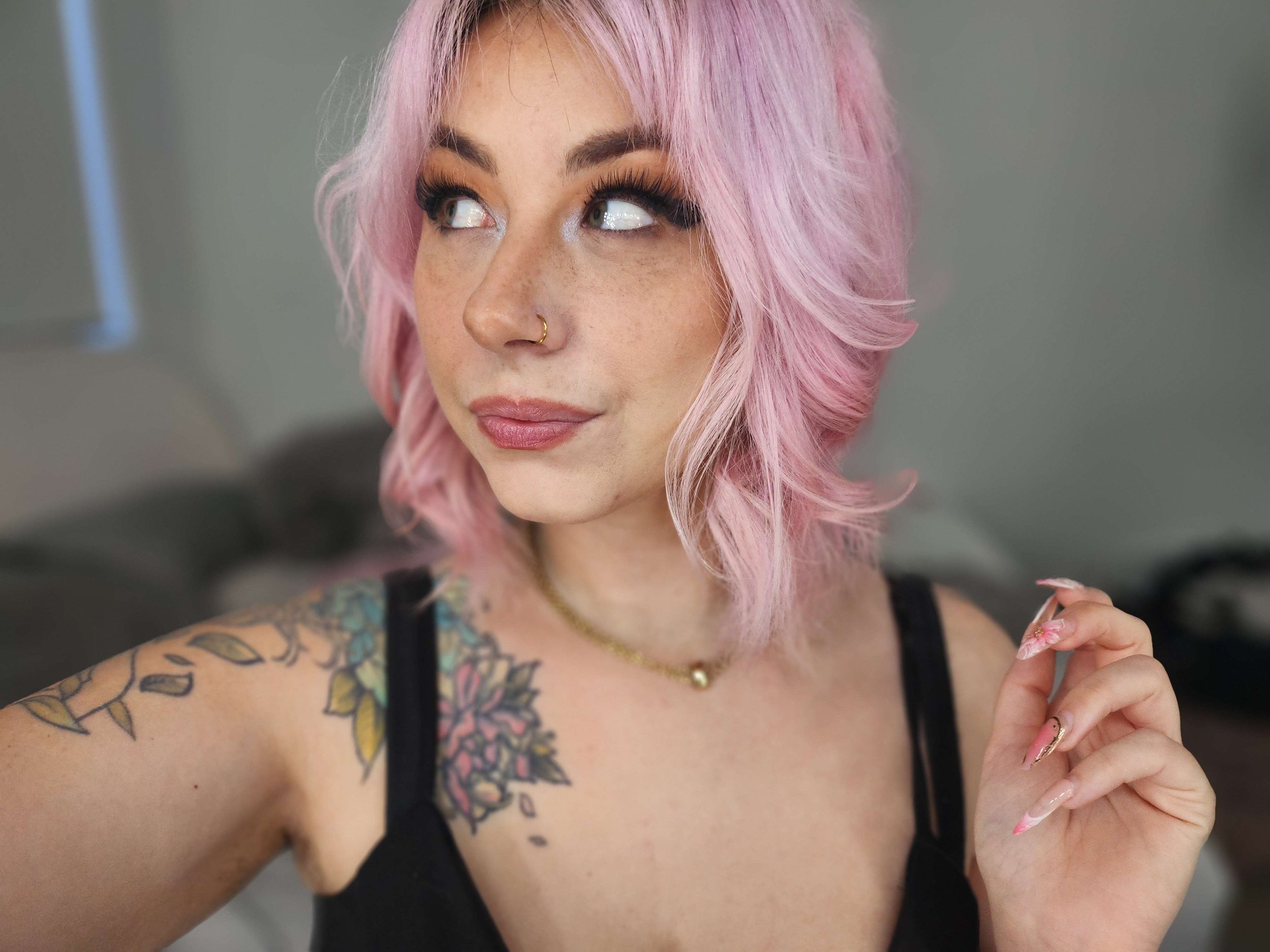 A woman with pink hair, freckles, a nose piercing, and tattoos on her arm and shoulder, wearing a black top and a gold necklace with a pearl pendant, looking to the side.