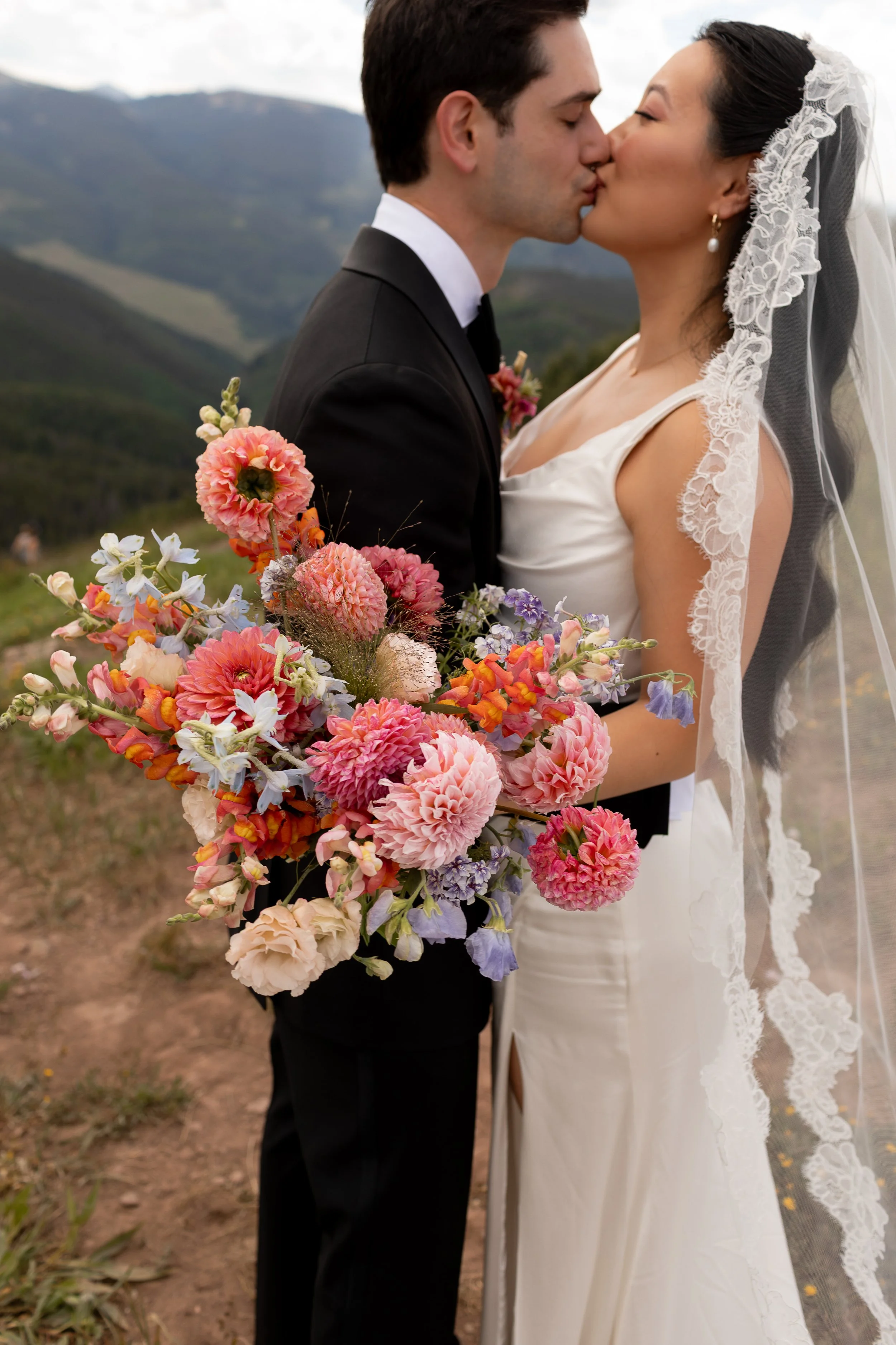 A bride and groom kiss outdoors, with the bride holding a colorful bouquet of pink, purple, and orange flowers, and mountains in the background.