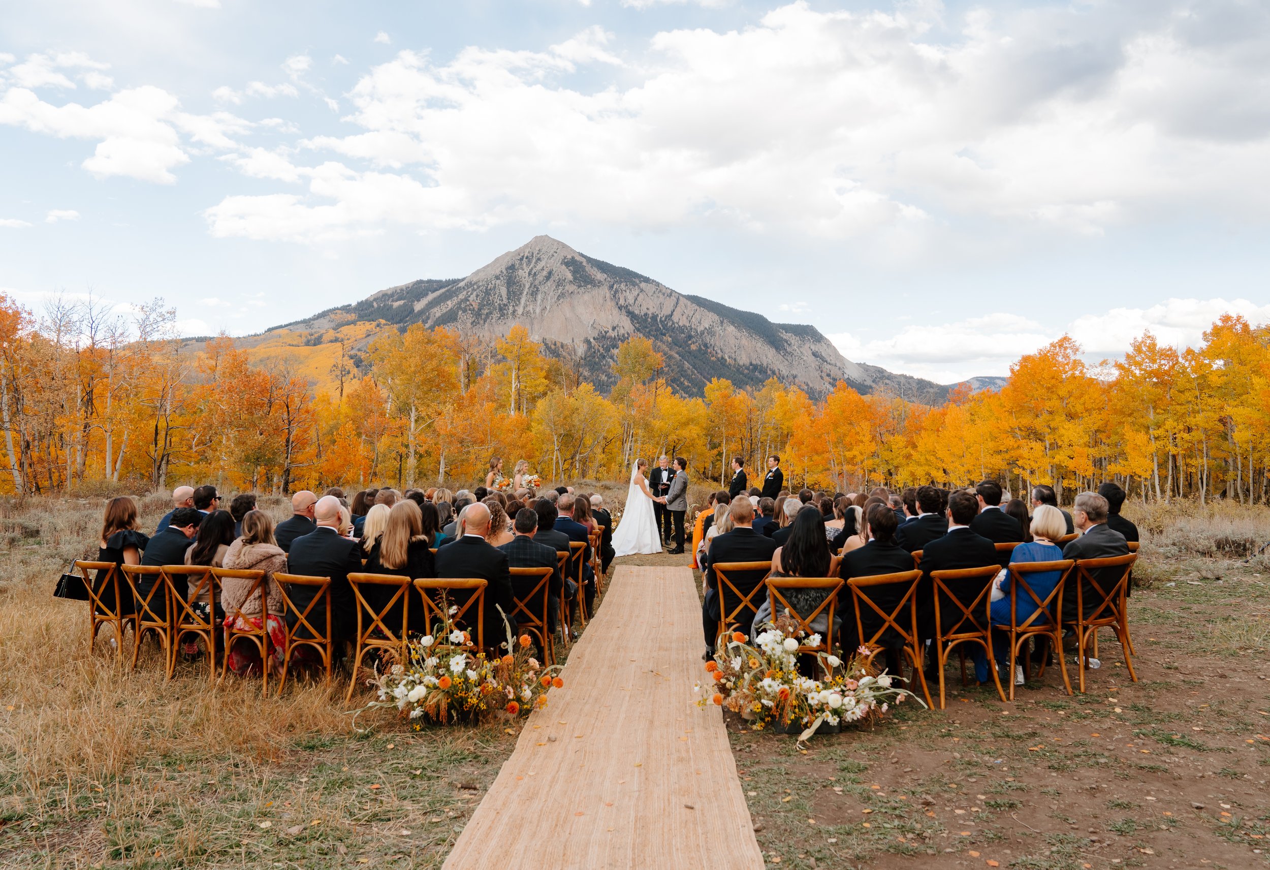 A bride and groom kiss outdoors, with the bride holding a colorful bouquet of pink, purple, and orange flowers, and mountains in the background.