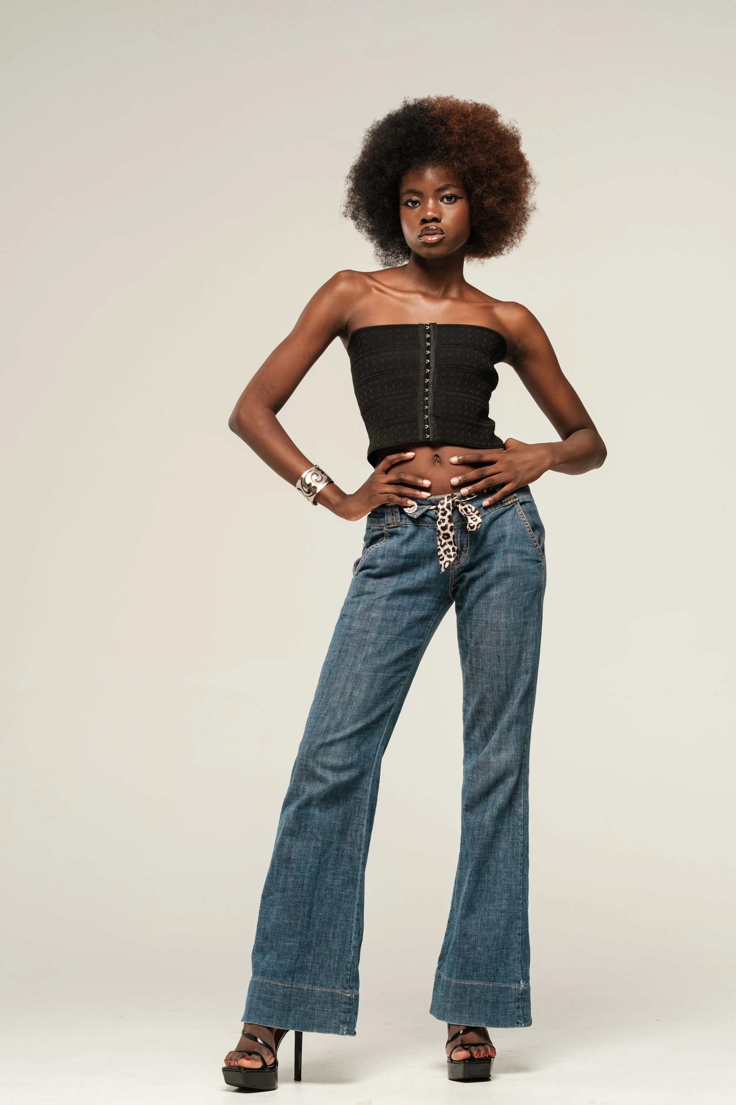 A woman with a large, natural afro hairstyle, wearing a strapless black top, blue jeans with a leopard print belt, black high-heeled sandals, and silver jewelry, standing with her hands on her hips against a plain background.