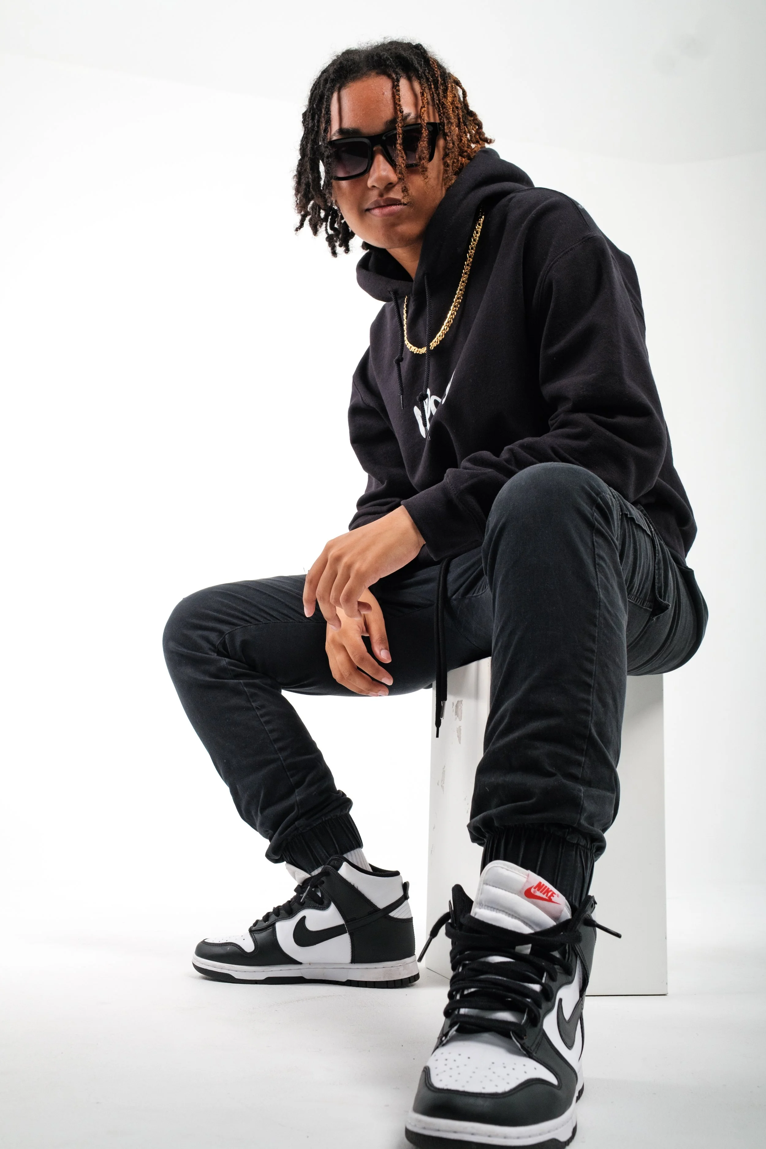 A young man wearing sunglasses, a black hoodie, gold chain, black joggers, and black-and-white Nike sneakers, sitting on a white cube against a white background.