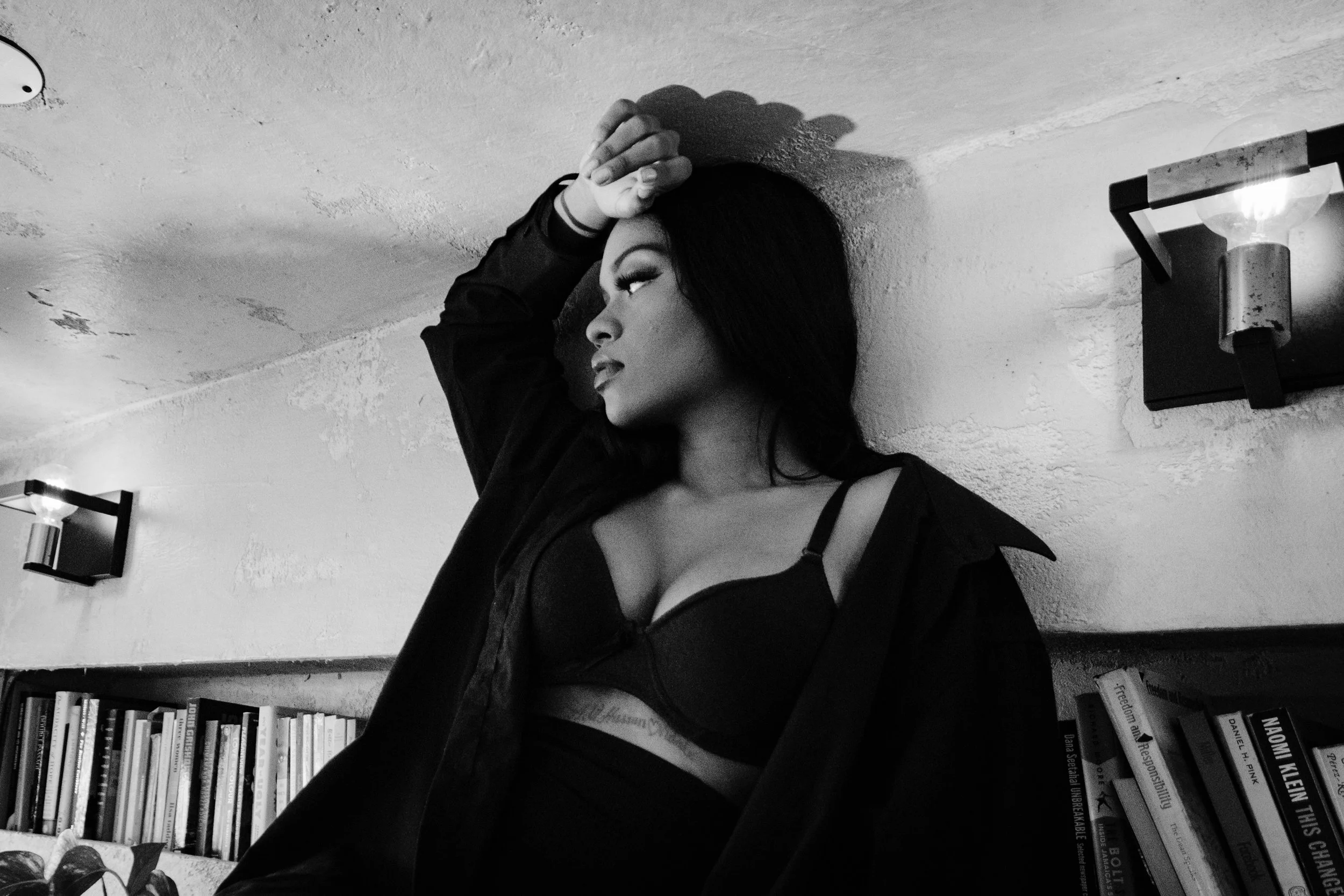 A woman with dark hair and makeup resting her head against a textured wall, wearing a black bra and a dark shirt, in a room with books and wall sconces.