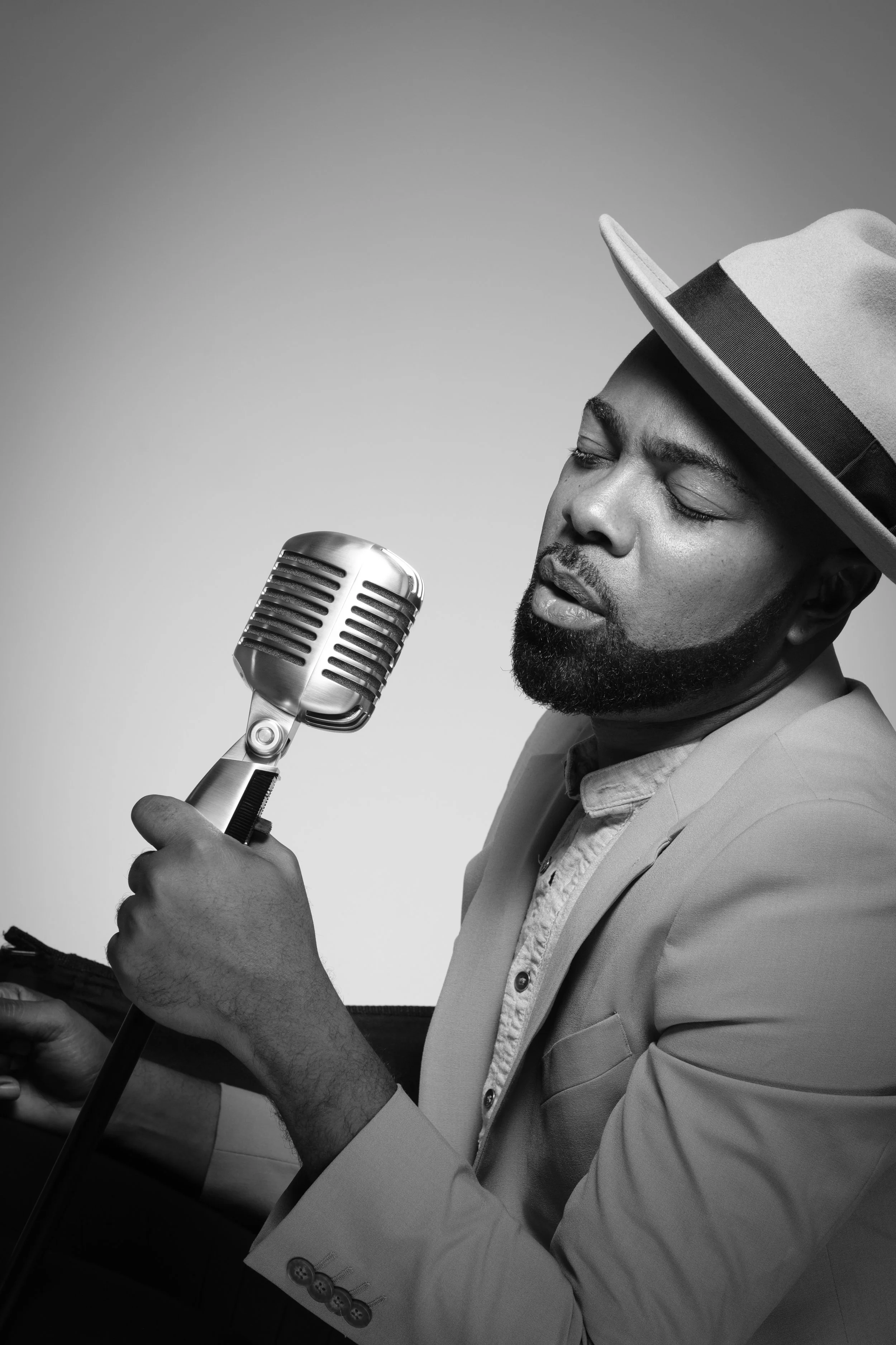 A man singing into a vintage-style microphone, wearing a light-colored blazer, a shirt, and a hat, with his eyes closed in a passionate performance.
