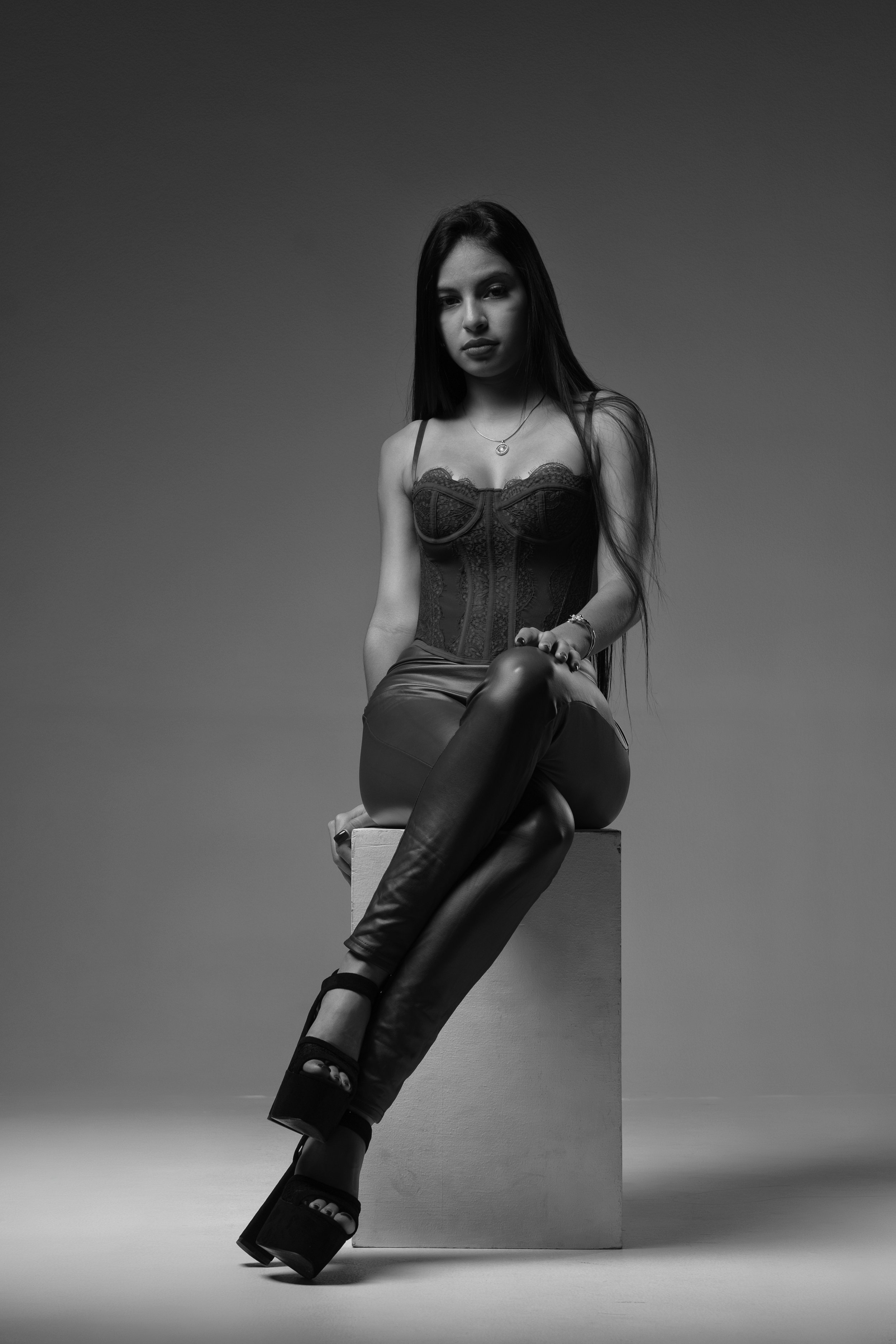 Black and white photo of a young woman with long hair, wearing a corset top, leather pants, and high platform shoes, sitting on a cube against a plain background.