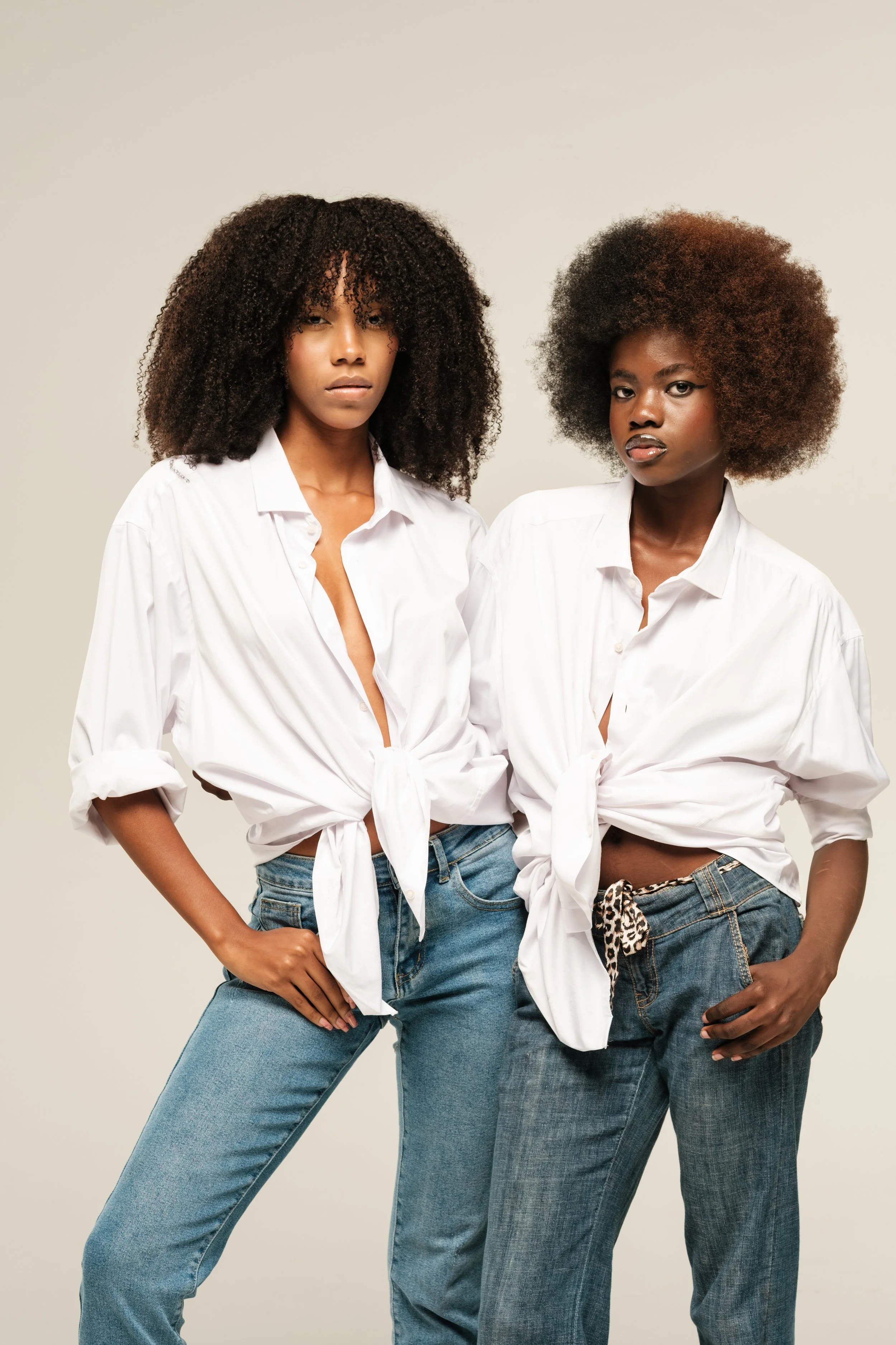 Two women with curly hair wearing white button-up shirts tied at the waist and jeans standing against a plain background.