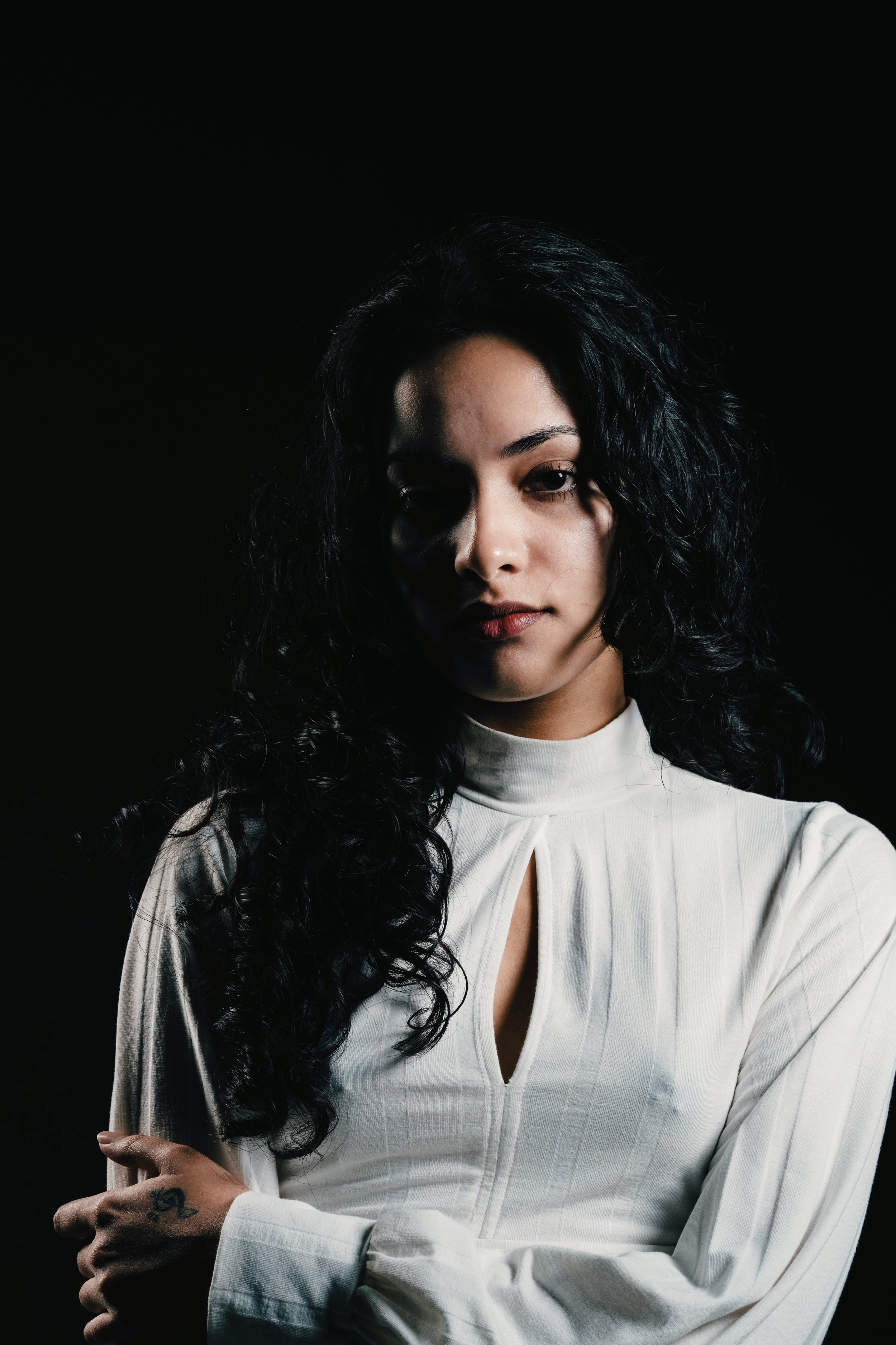 Portrait of a woman with dark, wavy hair wearing a white blouse with a keyhole neckline, cross-armed, against a black background.