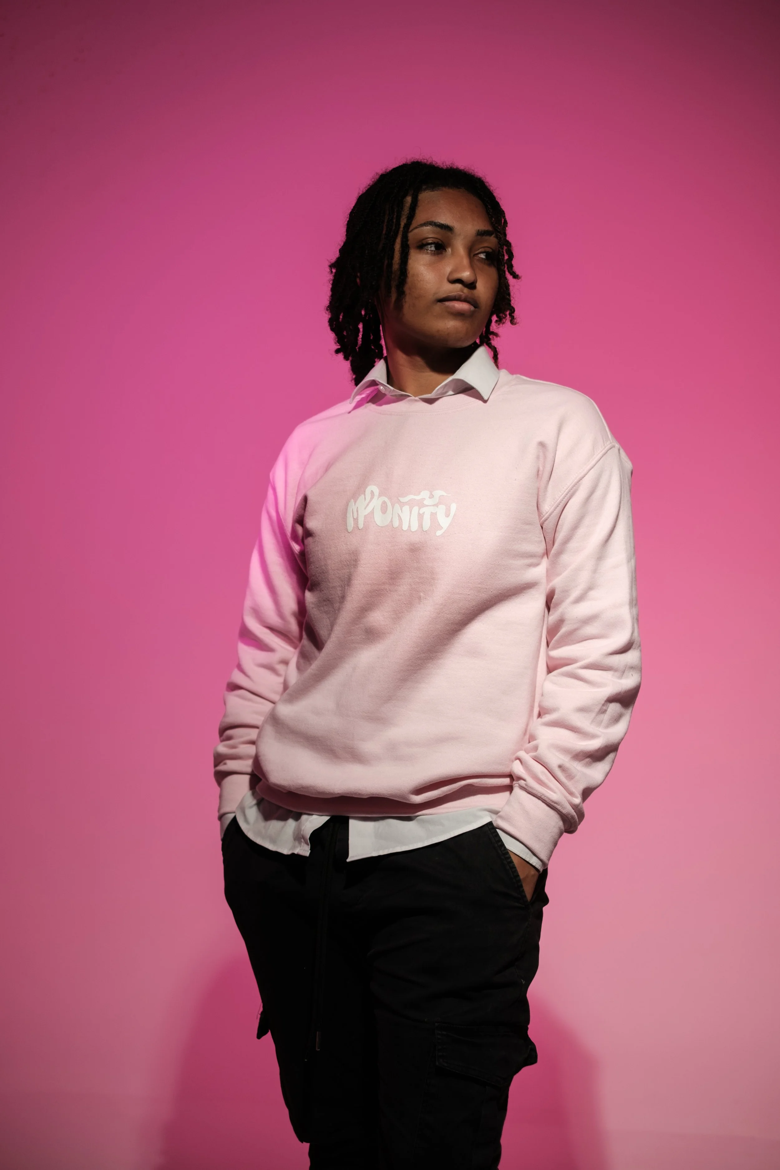 A person with dark curly hair wearing a pink sweatshirt with white text, standing against a pink background.