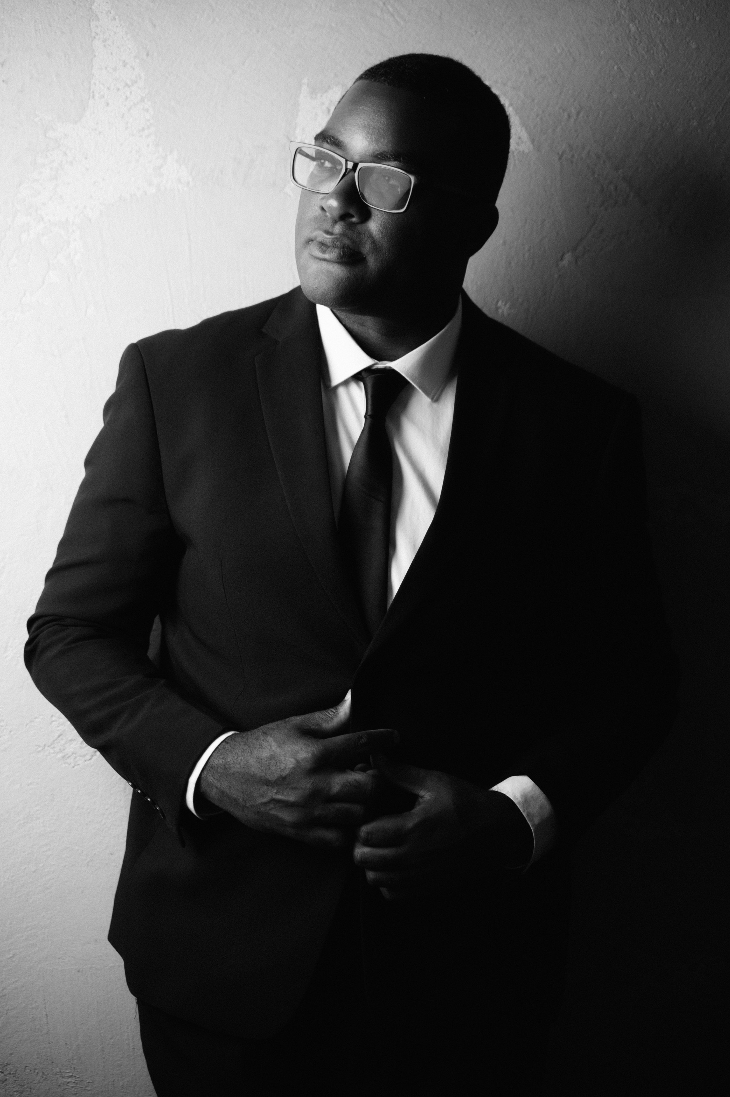 A man wearing a suit, tie, and glasses standing against a wall, looking to the side in black and white.