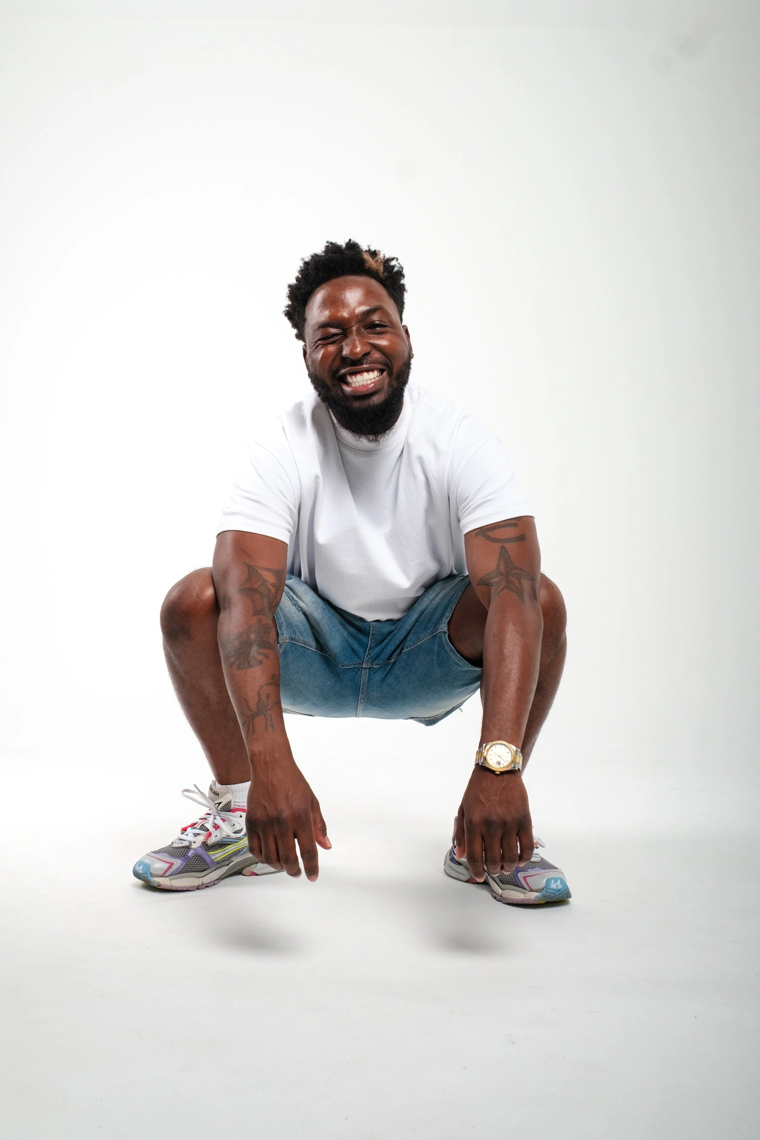 A man squatting in a white t-shirt, denim shorts, and sneakers, smiling at the camera against a plain white background.