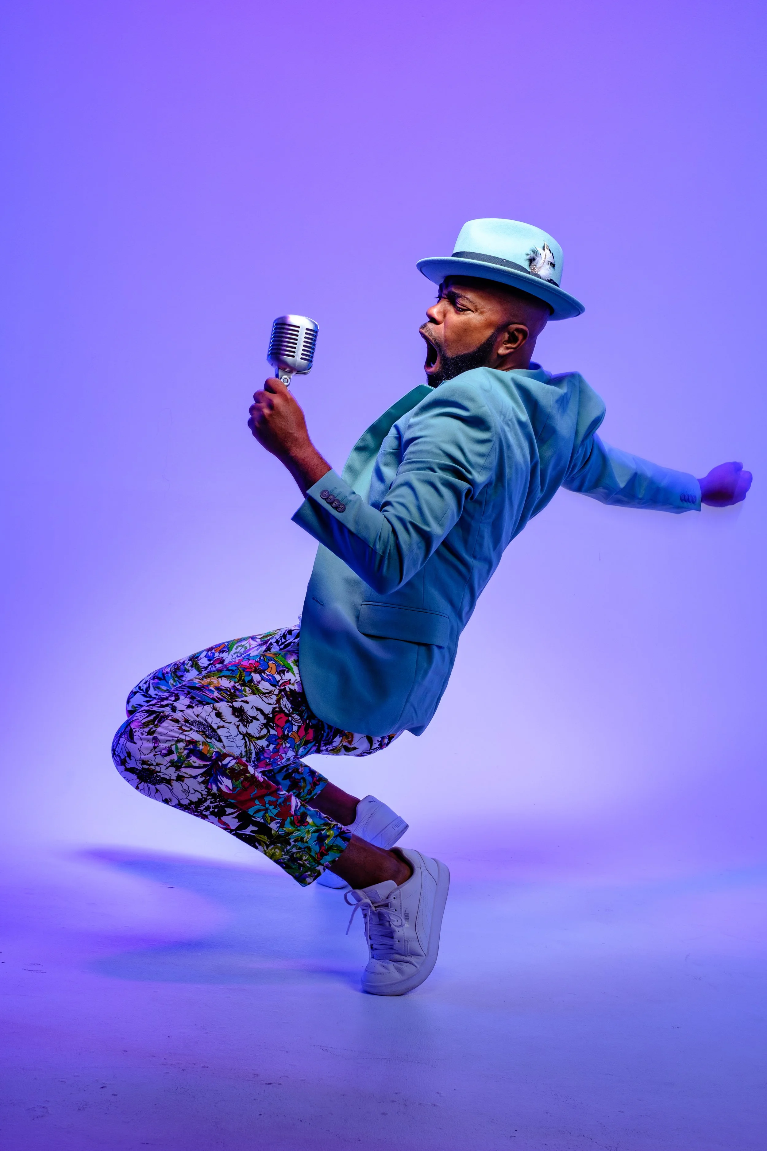 A man wearing a light blue jacket, patterned pants, white sneakers, and a light blue hat with a feather, singing into a vintage microphone with a purple background.