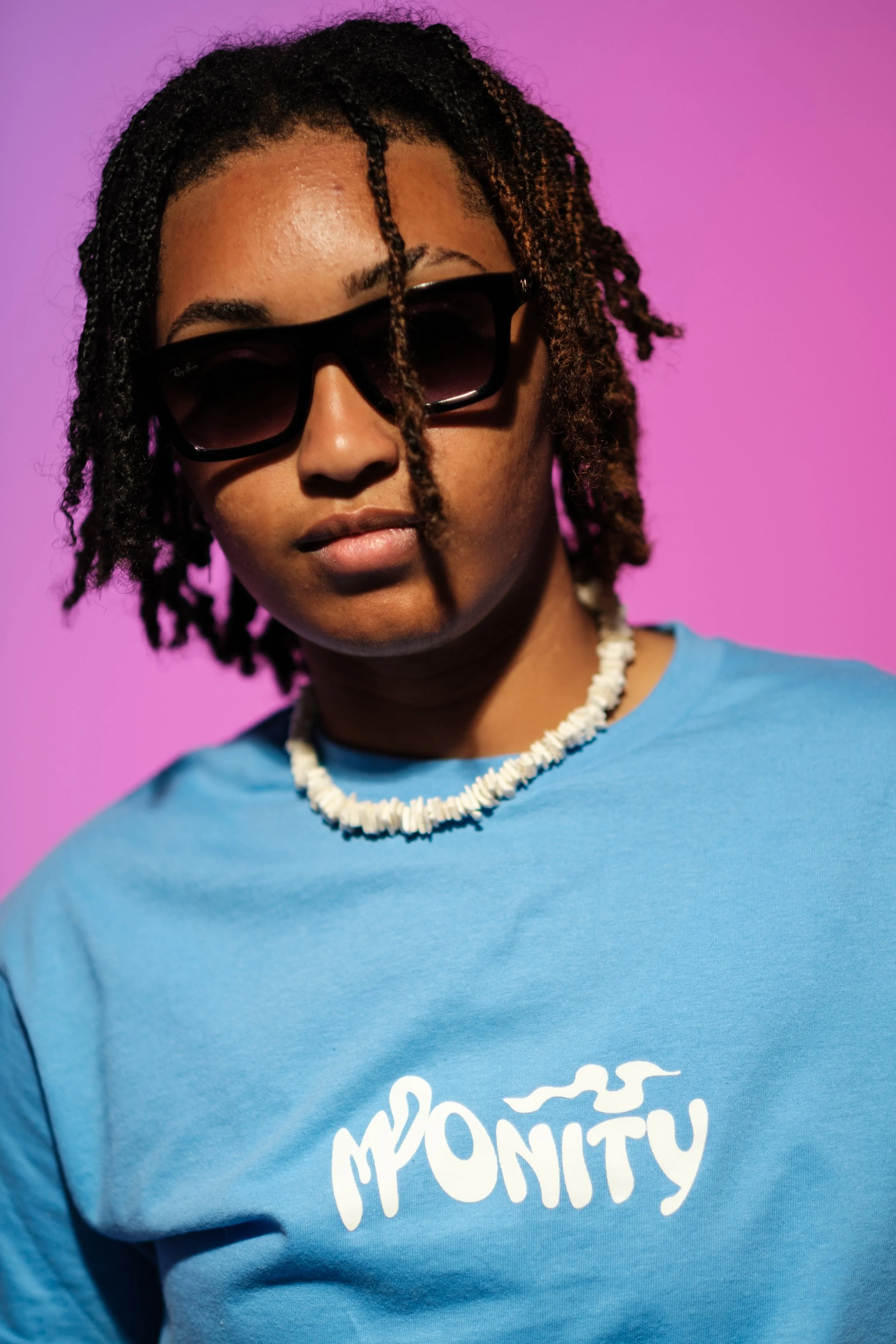 A woman wearing black sunglasses, a blue T-shirt with white text that reads 'MOMENTY', and a white beaded necklace, standing against a pink background.