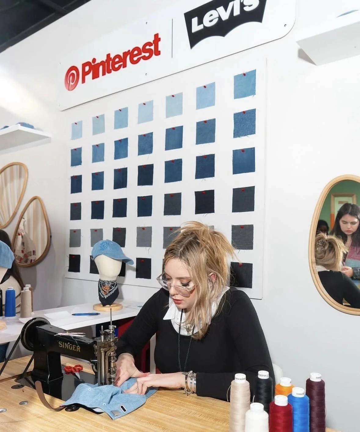 A woman sewing fabric on a black Singer sewing machine at a craft studio with Pinterest and Levi's branding on a wall behind her. There are spools of thread on the table and a mannequin wearing a blue cap and bandana. Reflection of people is visible 