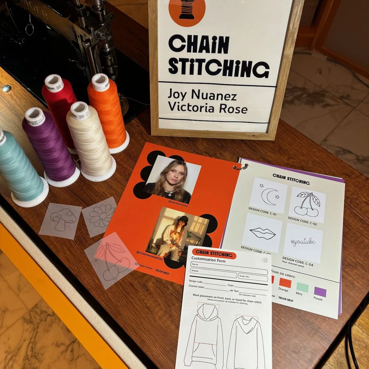 A table displays sewing supplies and promotional materials for a chain stitching workshop. There are spools of thread in purple, red, peach, orange, and mint colors, along with printed iron-on designs featuring a mushroom, flower, cherry, and 