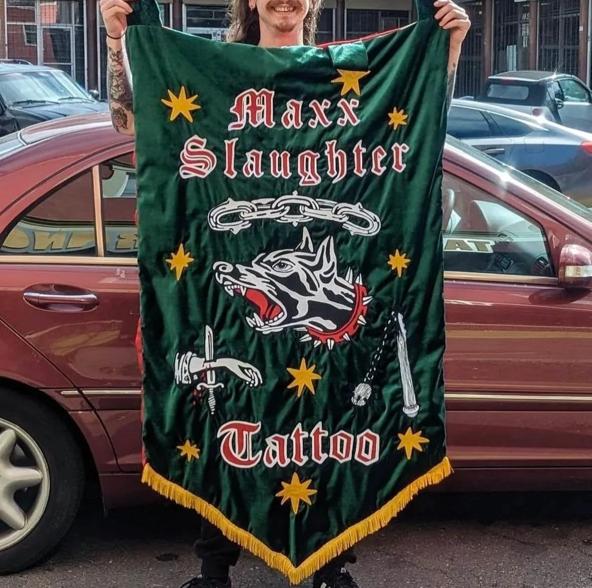 Person holding a green chainstitched banner that reads 'Maxx Slaughter Tattoo' with a snarling wolf head, chain, dagger, and stars, standing in front of a maroon car.