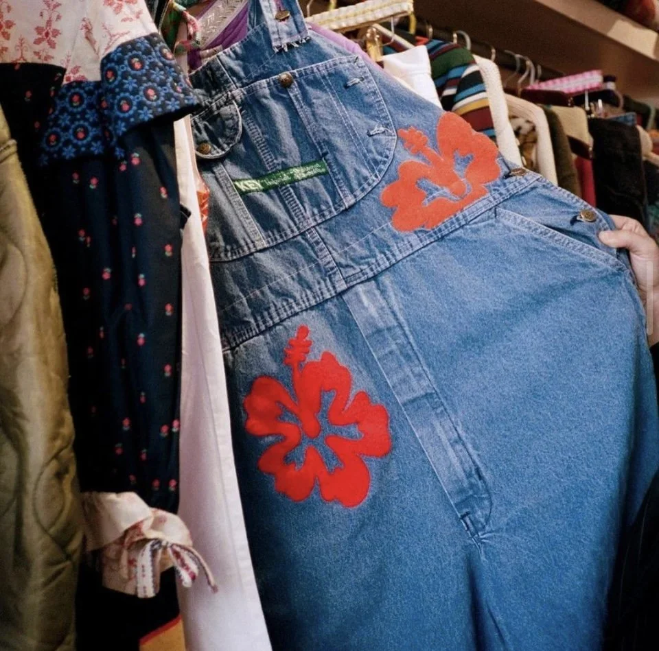 A rack of clothing featuring a pair of blue denim overalls with red floral embroidery on the back and a green tag, surrounded by various other garments in a thrift or vintage store.