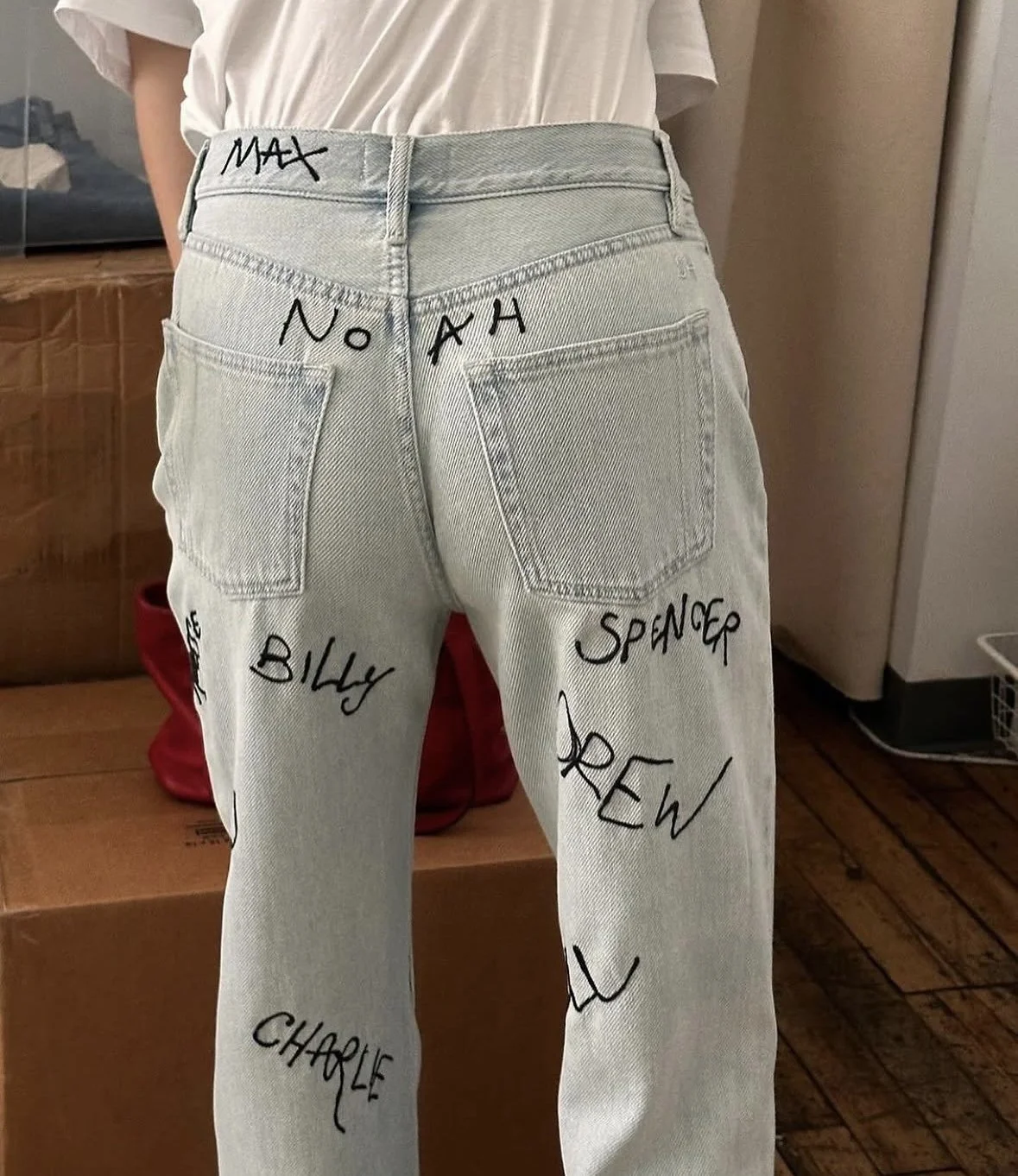 Back view of light-wash jeans with handwritten black names on them, including 'MAX', 'NOAH', 'BILLY', 'SPENCER', 'CREW', and 'CHARLIE', worn by a person in a white T-shirt.