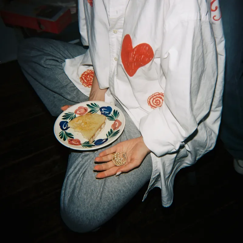 Person sitting on the floor holding a plate with a slice of pie. They are wearing a white jacket with a red heart and orange design. Their other hand is resting on their knee, holding a decorative ring. The person is dressed in gray sweatpants.