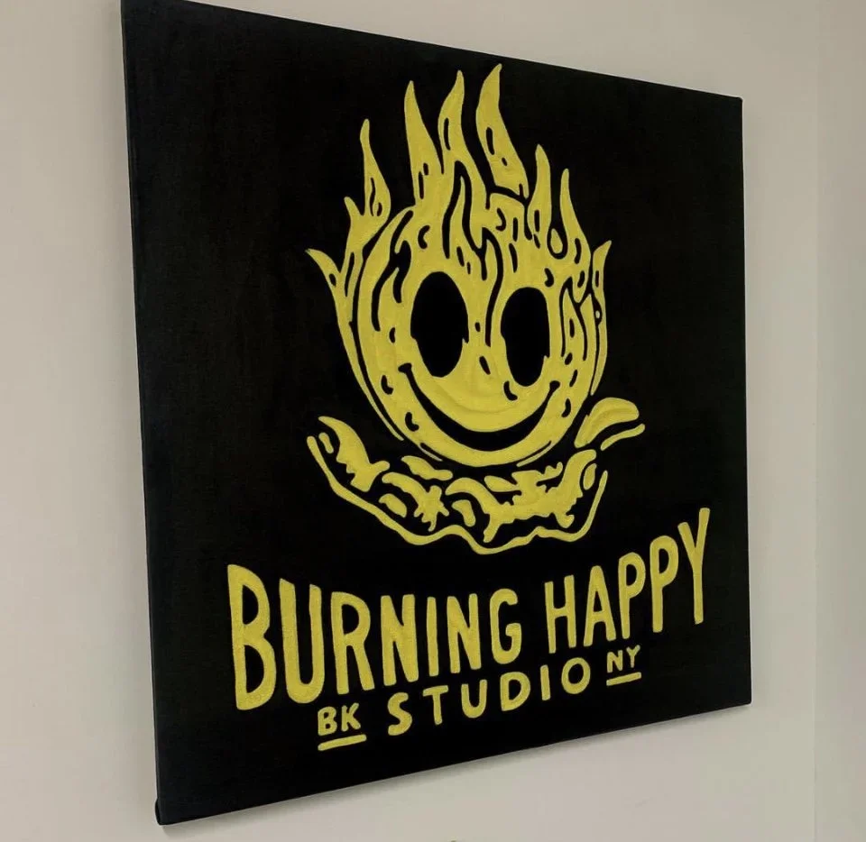 A black canvas with a yellow smiling flame logo and text that reads "BURNING HAPPY BK STUDIO NY."