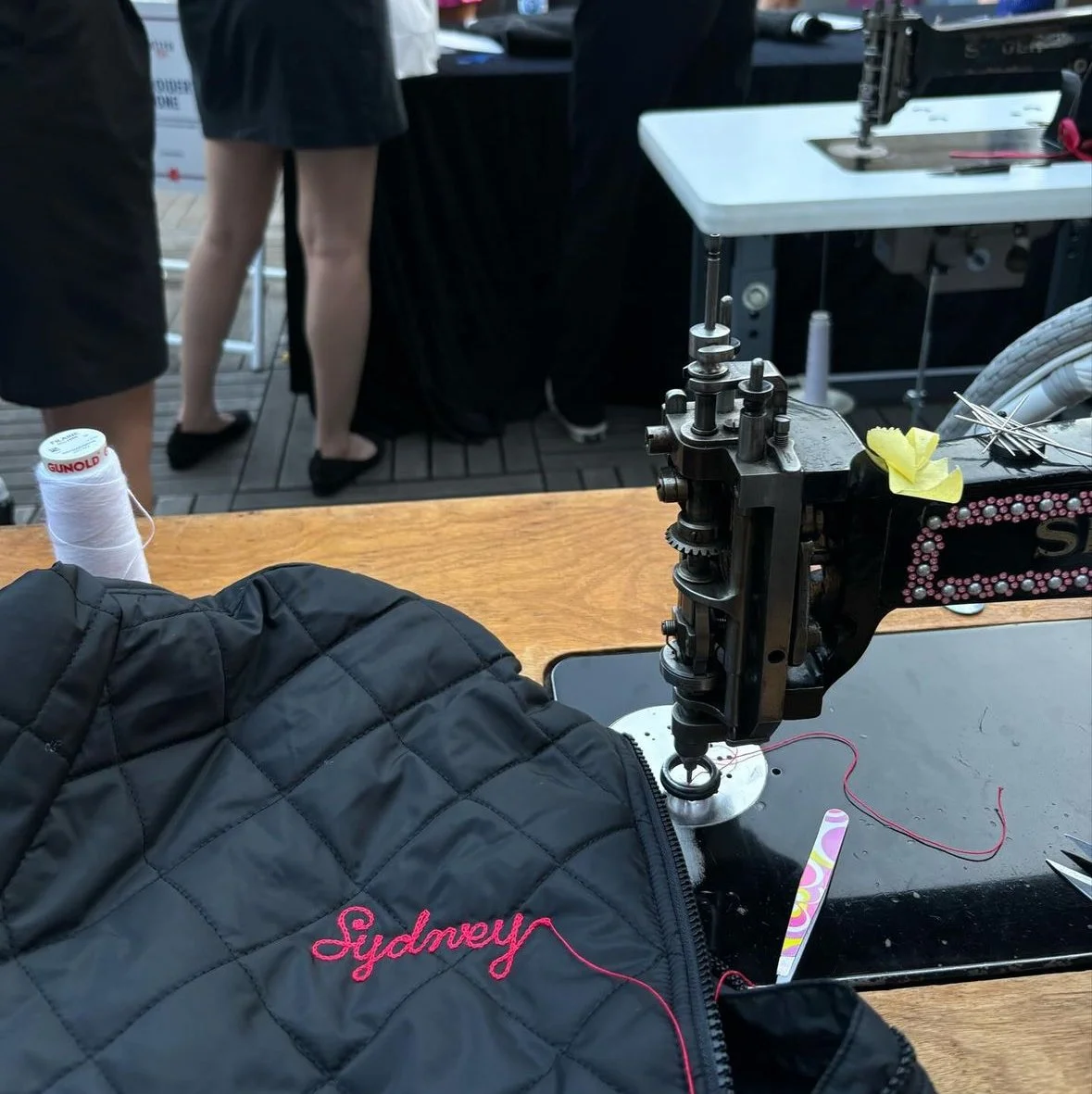 Close-up of an industrial sewing machine with colorful embellishments, on a wooden table, with a black quilted jacket with pink embroidered name 'Sydney' in the foreground, and people walking in the background on a wooden deck.