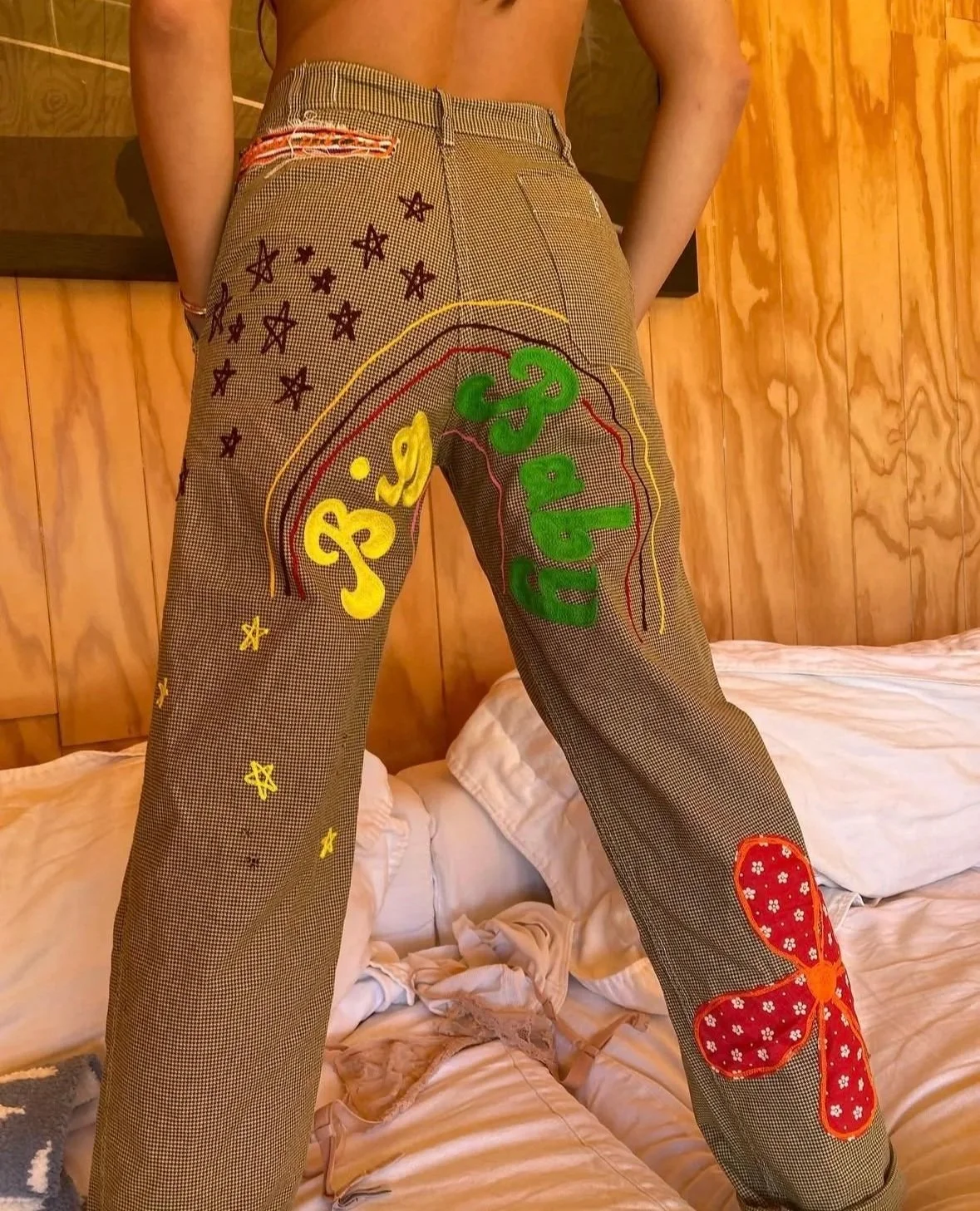 Person wearing pants with colorful embroidered designs and text, standing on a bed with wooden wall background.