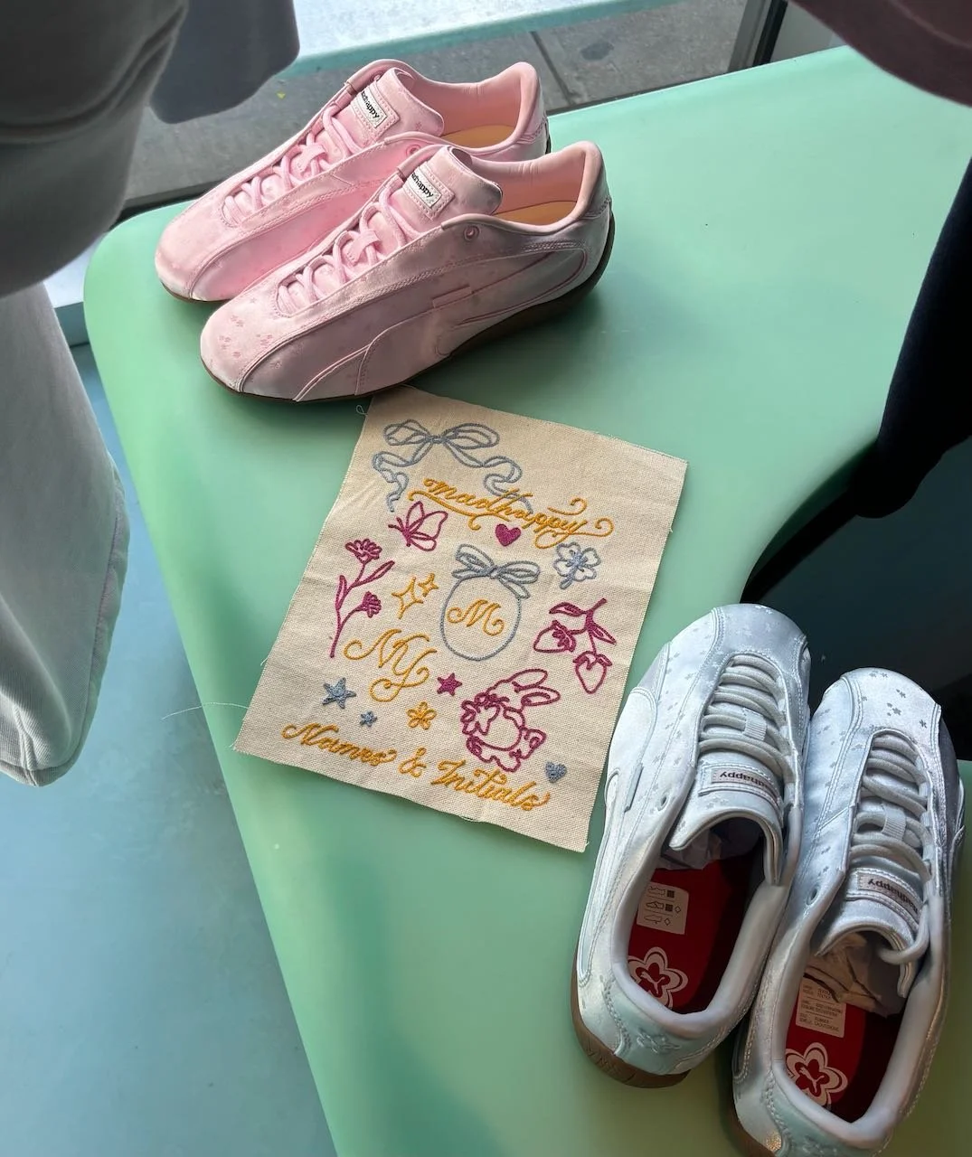 Pink sneakers on the left, white sneakers on the right, embroidered cloth with colorful designs and text, displayed on a green table.
