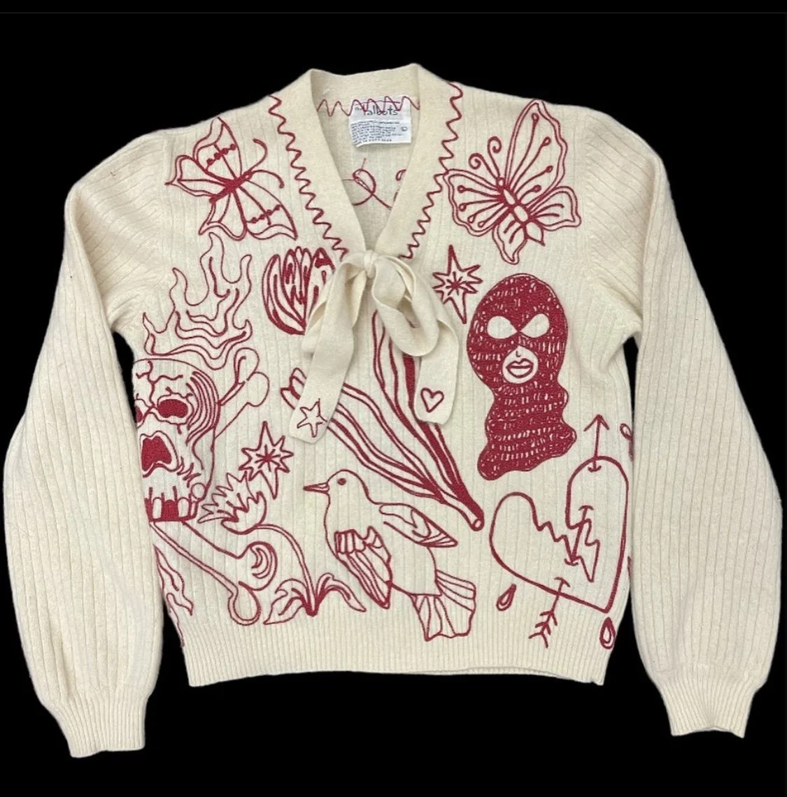 Cream-colored knitted sweater with red doodle-style illustrations and designs, including butterflies, a skull, a woman with a mask, a bird, stars, a heart, and various abstract patterns, with a ribbon tie at the neckline.