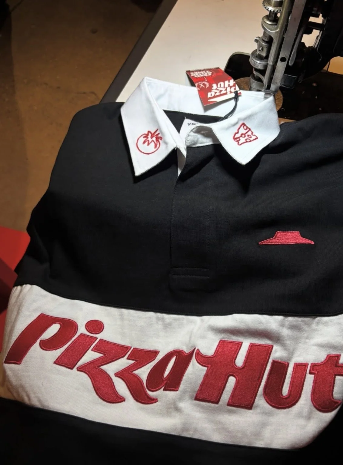 A stuffed toy in the shape of a little boy dressed in a black and white uniform resembling a Pizza Hut employee, sitting on a table, with a press machine and a Pizza Hut shopping bag nearby.
